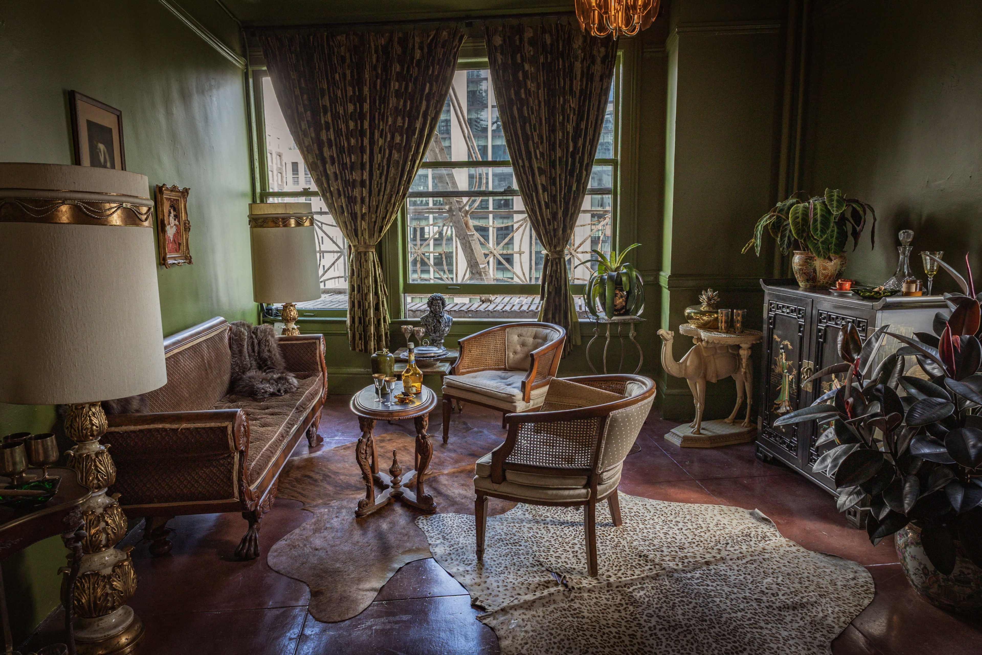 The image shows a vintage-style living room featuring a brown sofa, two armchairs, a small round table, and decorative plants, all set against green walls and large windows.