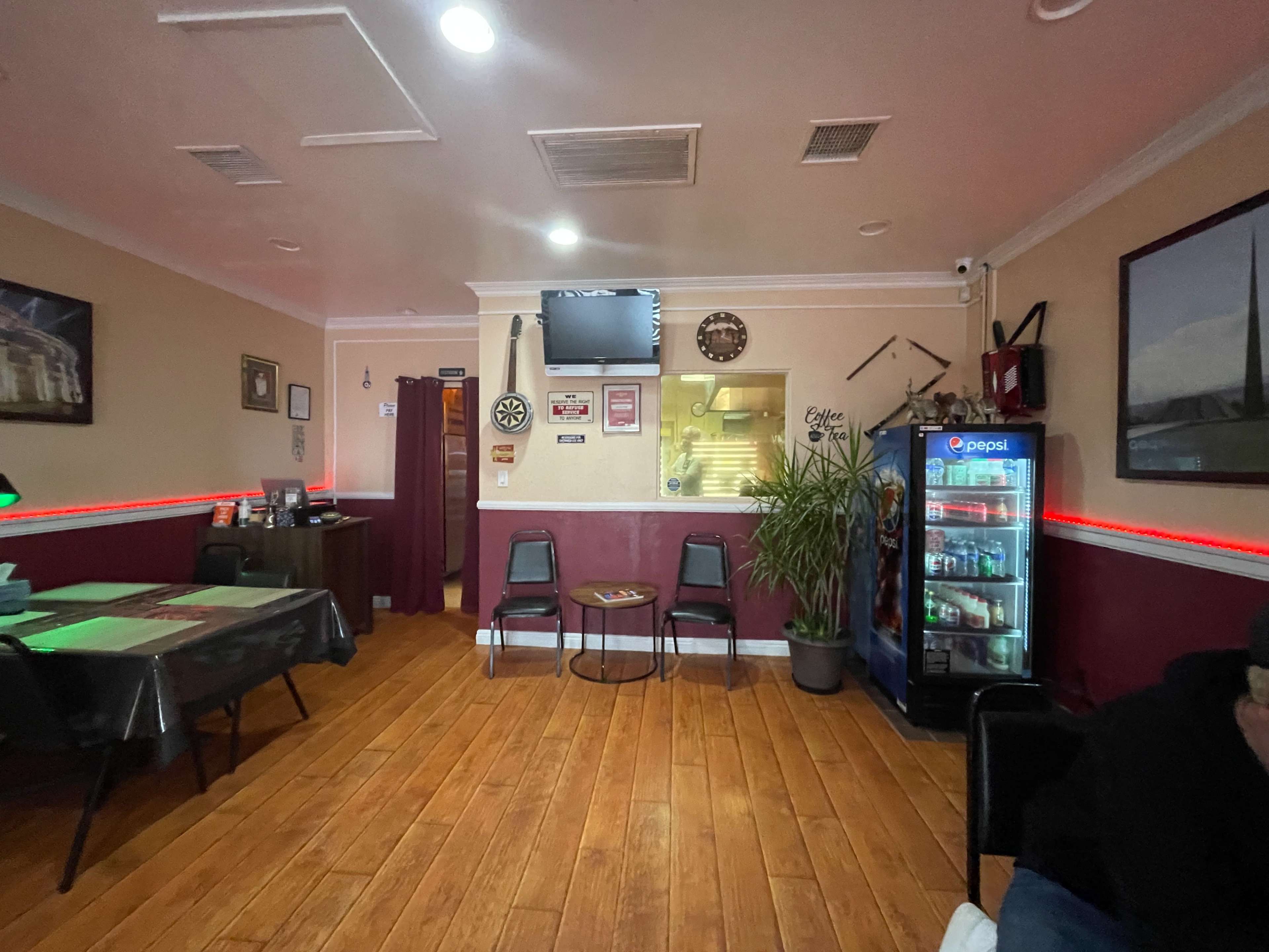 The image shows the interior of a small restaurant featuring a seating area with tables and chairs, a television on the wall, and a plant next to a beverage cooler.