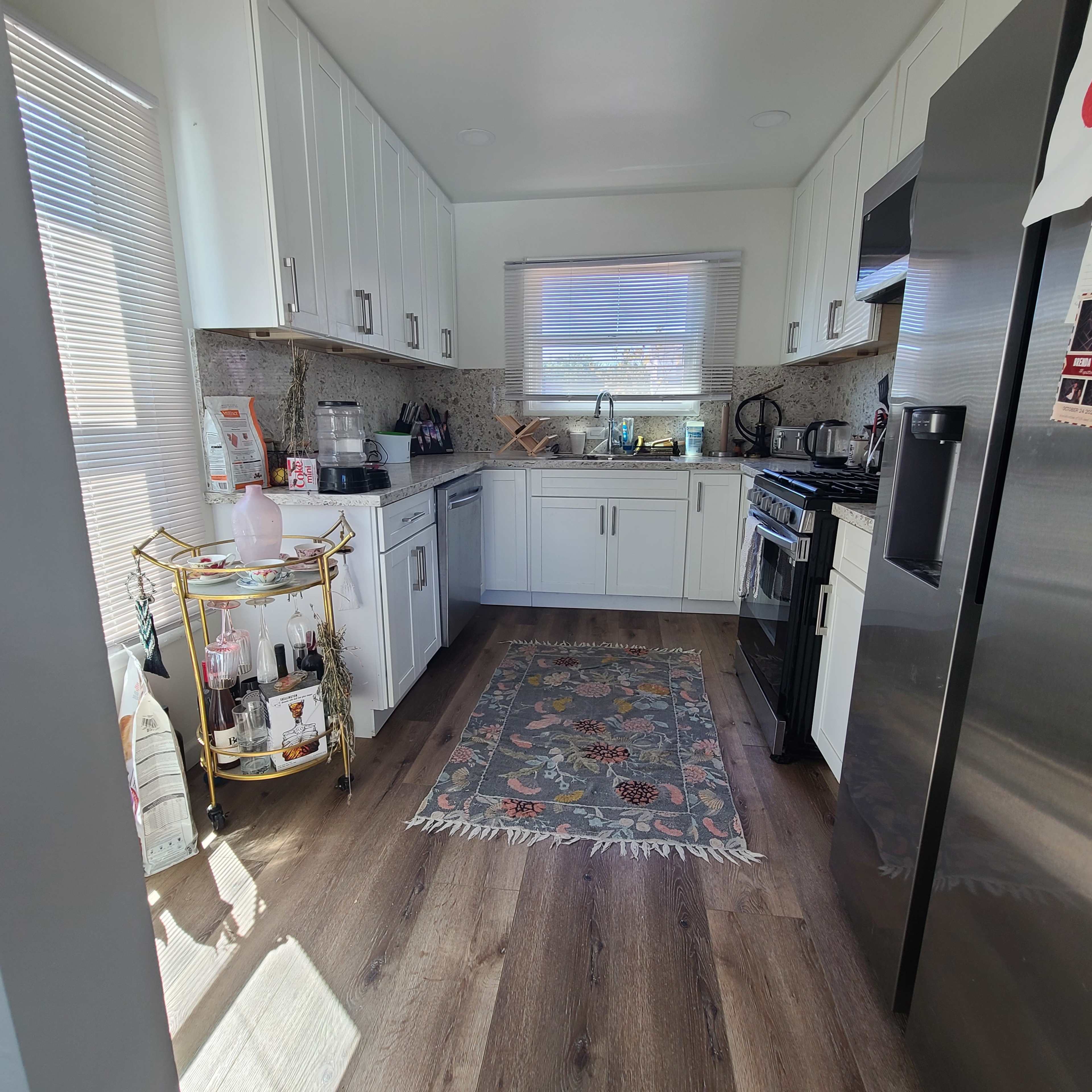 The image shows a modern kitchen with white cabinetry, a stainless steel refrigerator, and a decorative rug on the wooden floor.