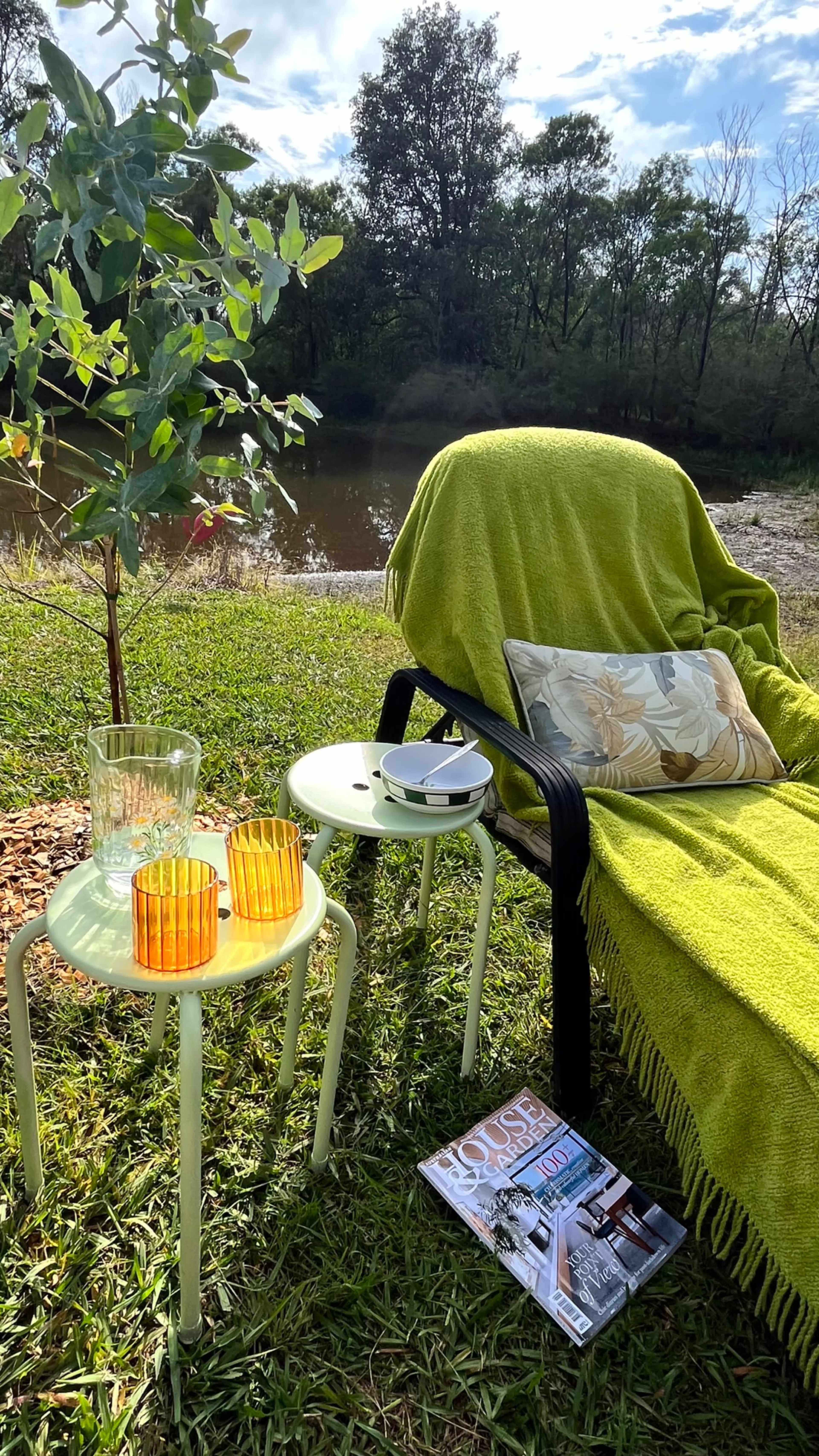 A chair draped with a green blanket and a cushion sits beside two small tables with drinks and a magazine, overlooking a riverbank surrounded by trees.