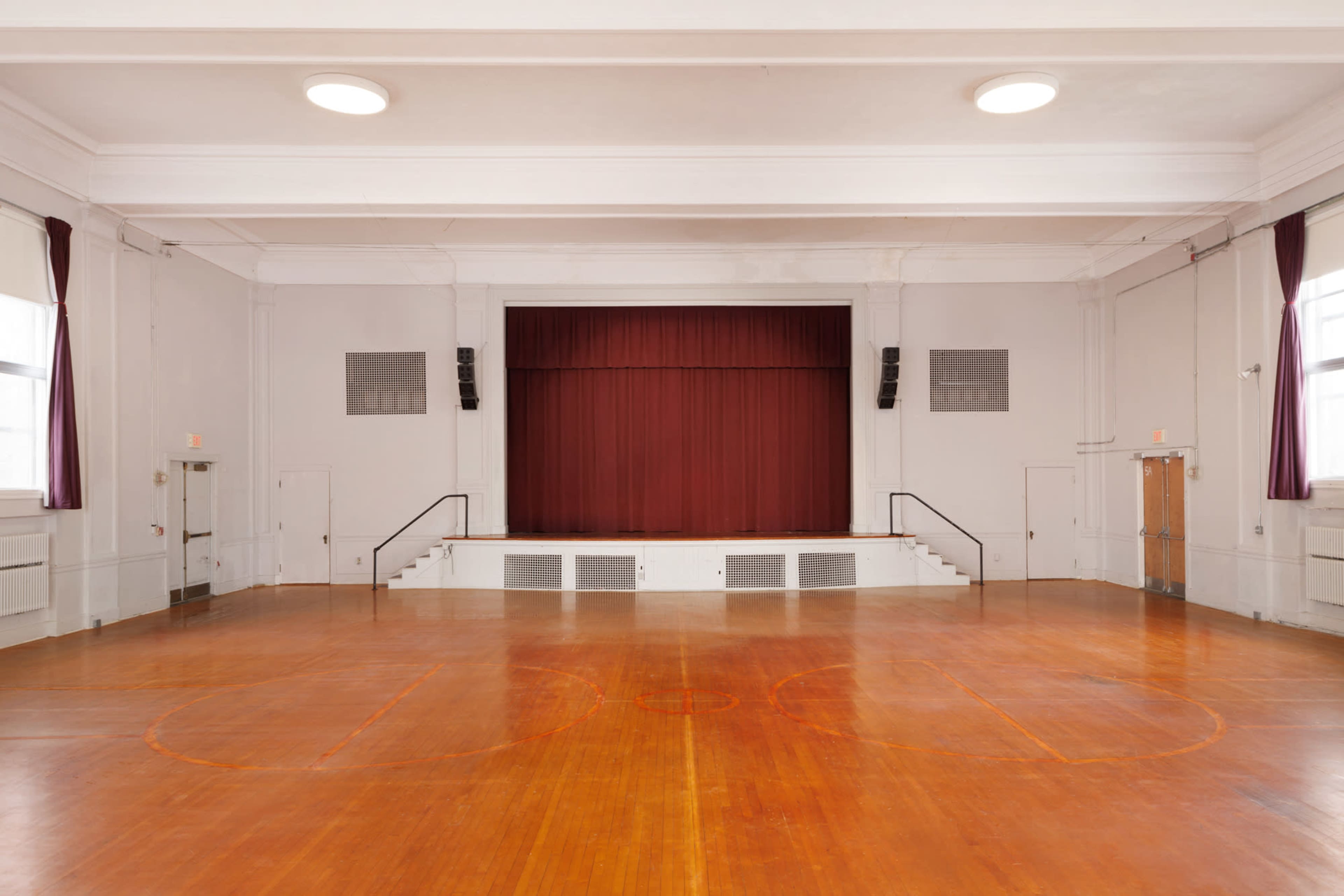 An empty gymnasium with a wooden floor and a stage at the front covered by a red curtain.