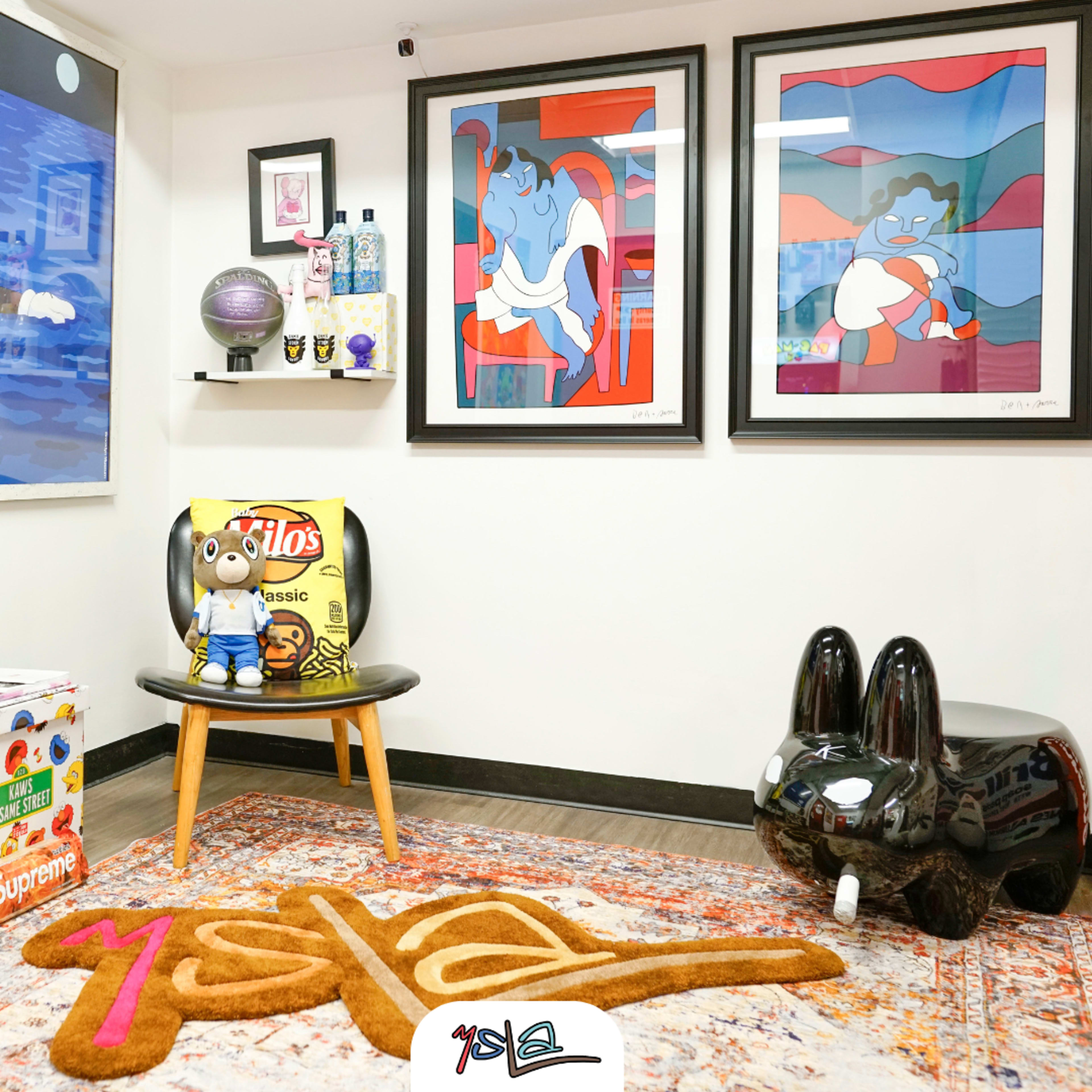 A modern room features a wooden chair with a cartoon-themed cushion, a small black sculpture, colorful artwork on the walls, and a patterned rug with the word "m̶e̶s̶t̶a̶l."