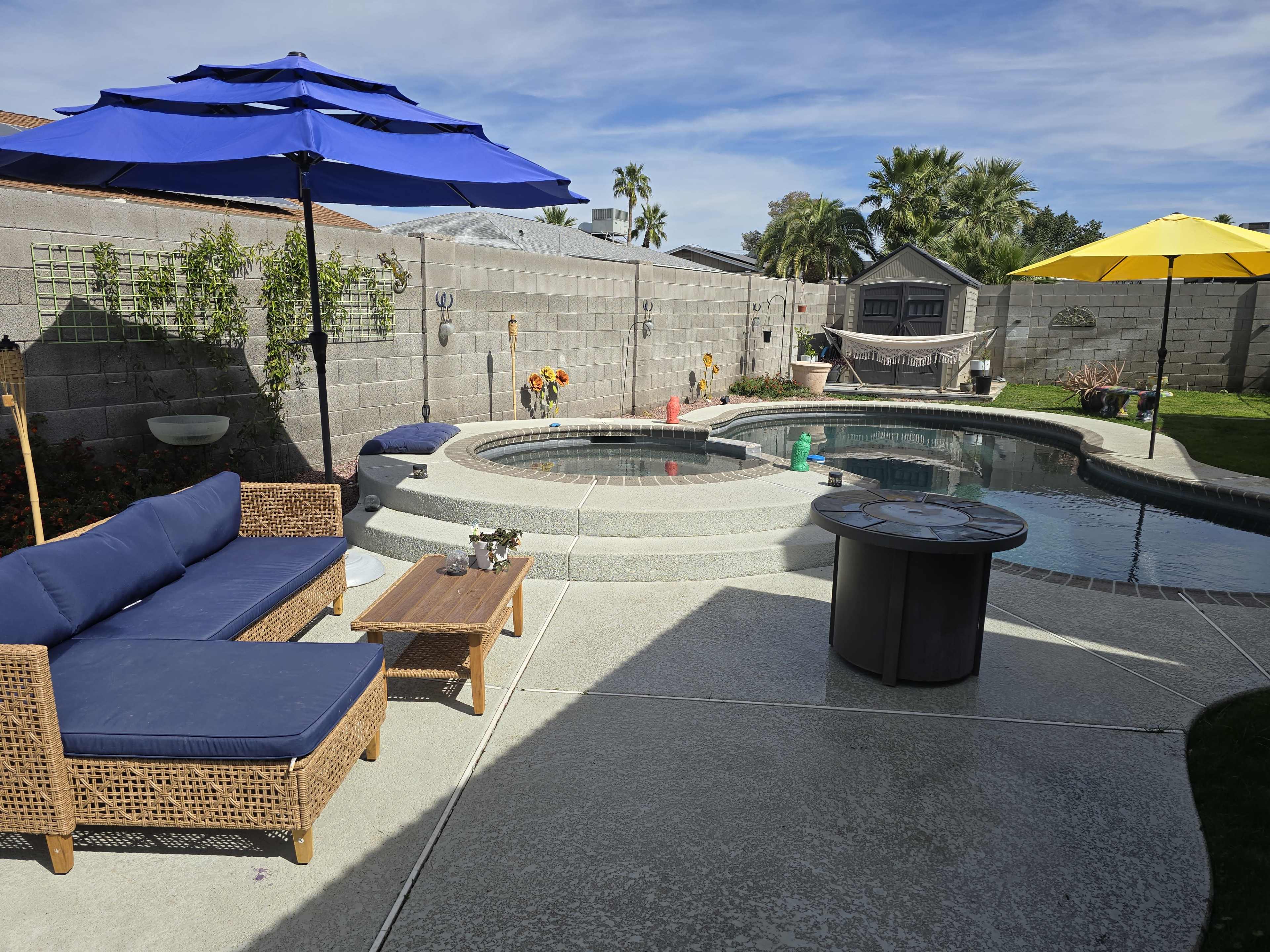 The image shows a backyard pool area with a circular spa, patio furniture, and two umbrellas—one blue and one yellow—set against a concrete wall.