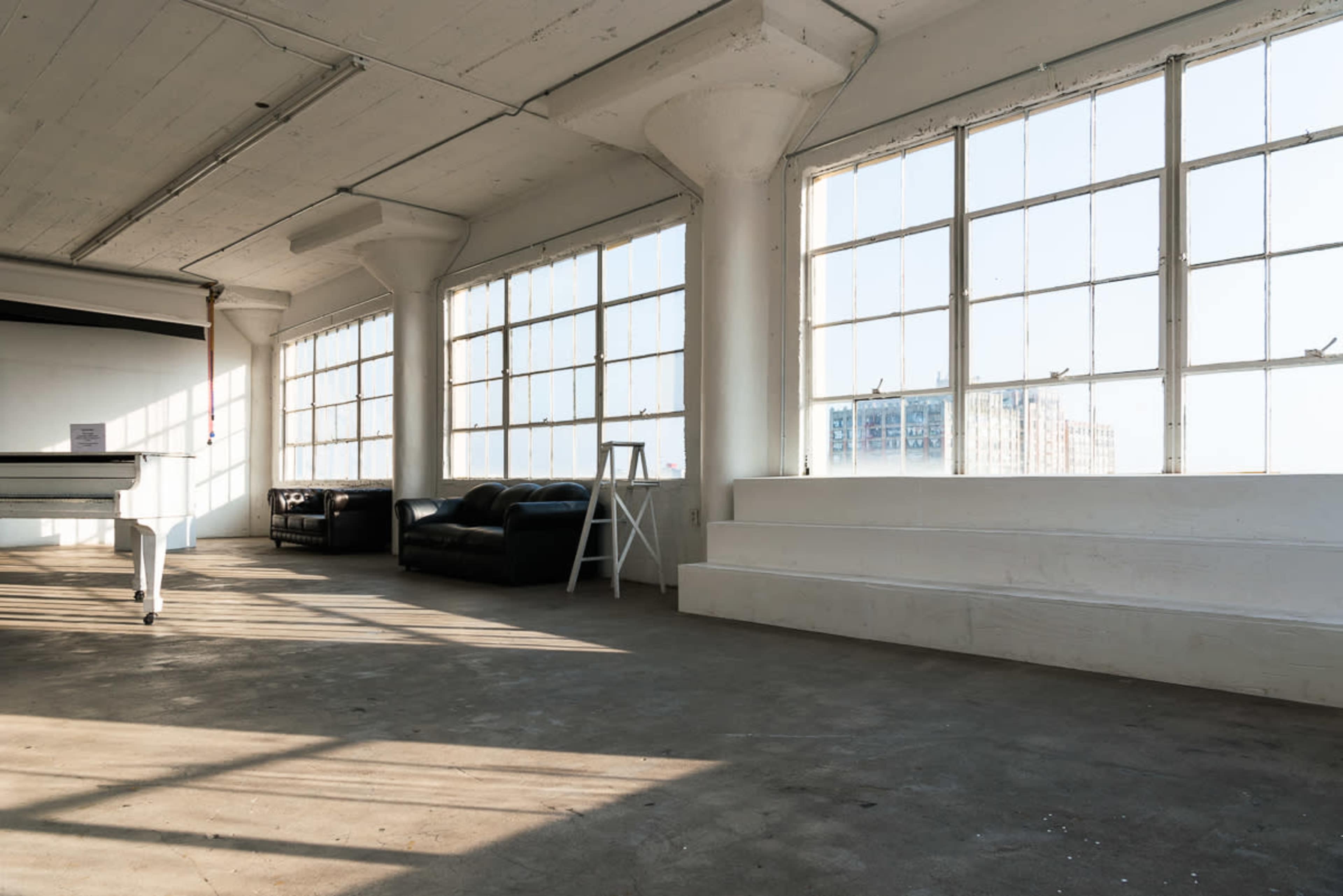 The image shows a spacious, well-lit interior featuring large windows, a black leather couch, and a staircase with a white wall background.