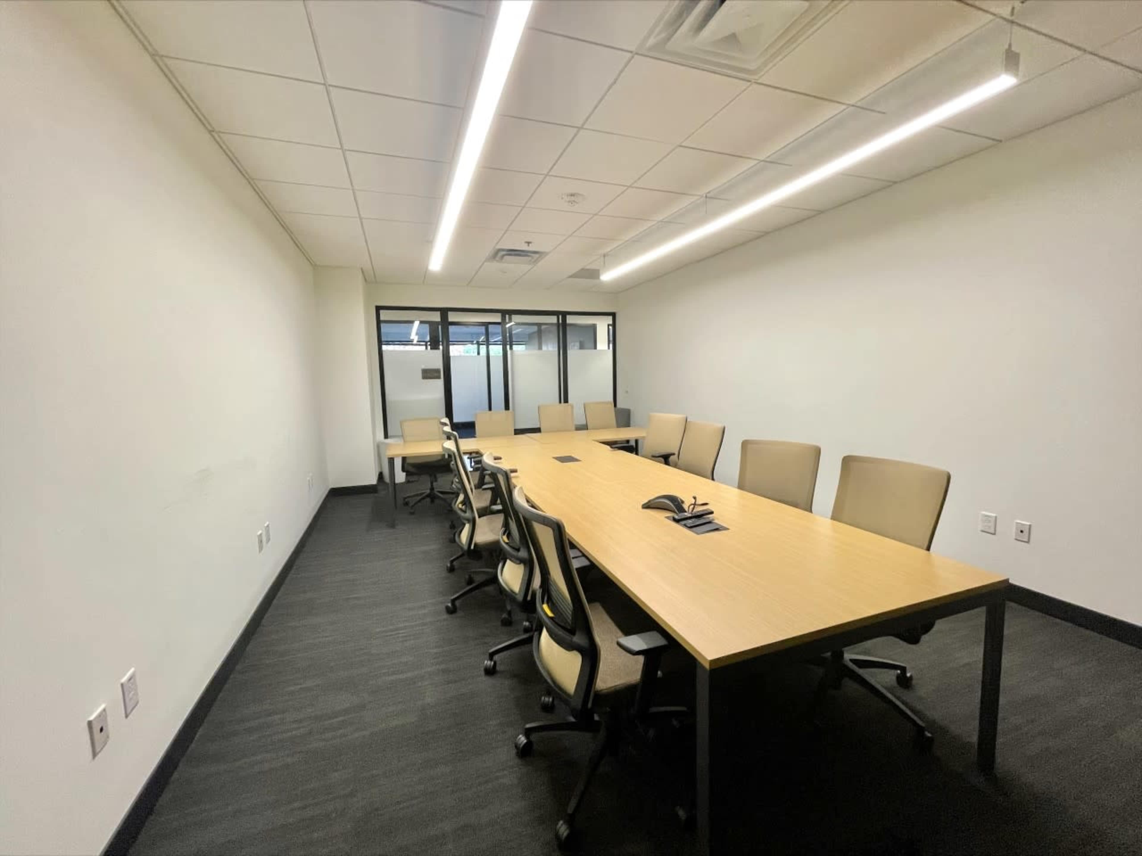A long conference table with chairs is placed in a well-lit meeting room featuring white walls and a floor covered in dark carpeting.