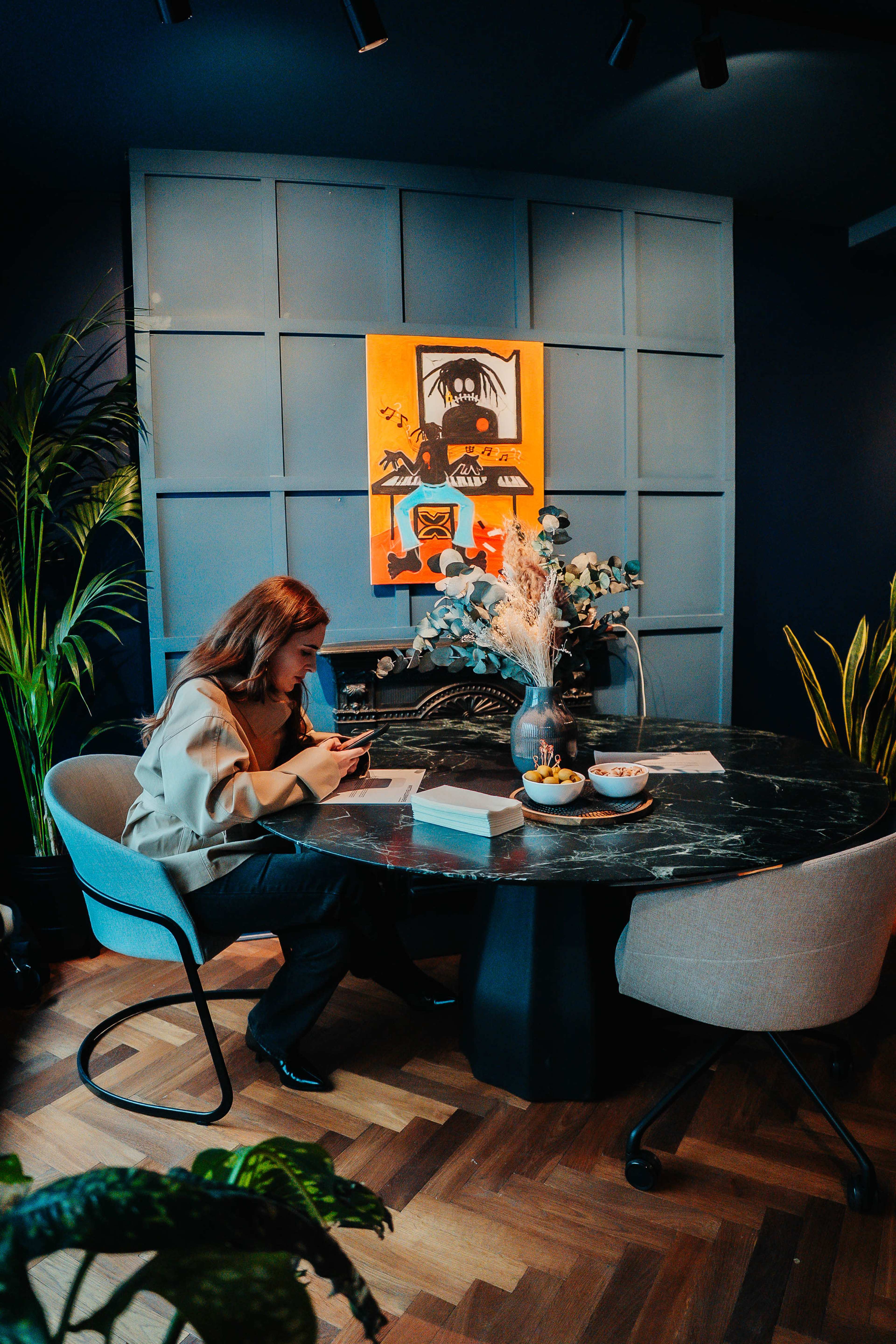 A woman sits at a round table in a stylish office space, using her phone while surrounded by plants and a colorful painting on the wall.