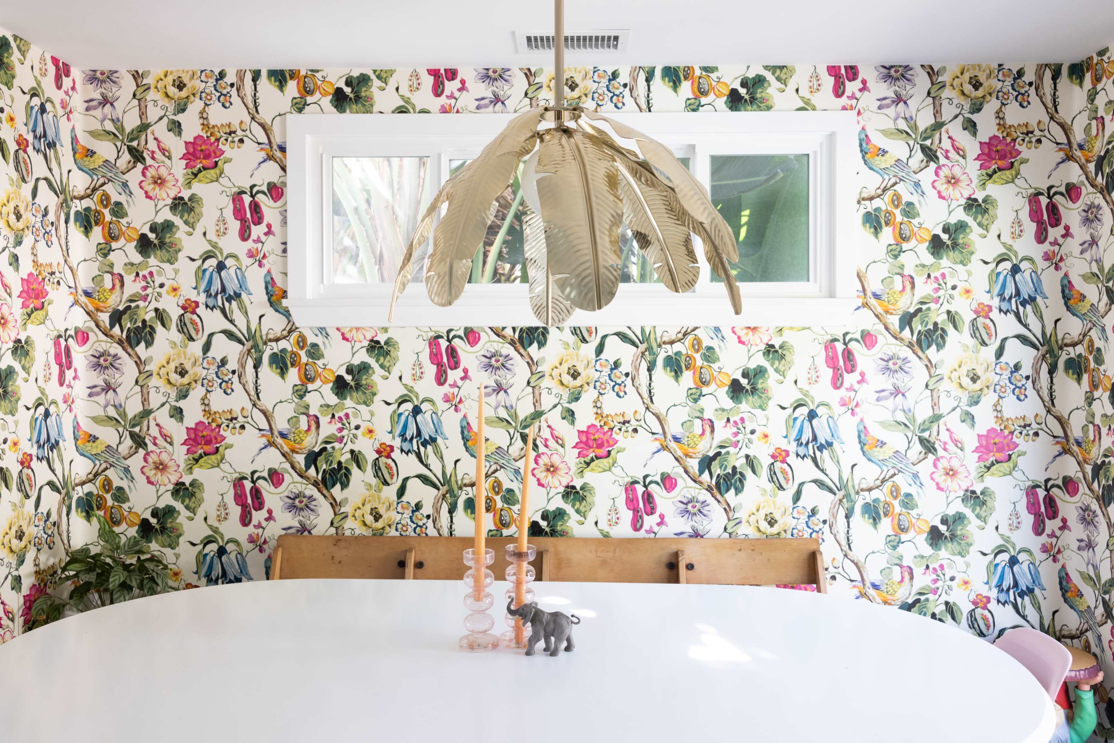 A round white dining table is set in a brightly decorated room with floral wallpaper, featuring a feather-style chandelier and decorative items like candles and a small elephant figurine.