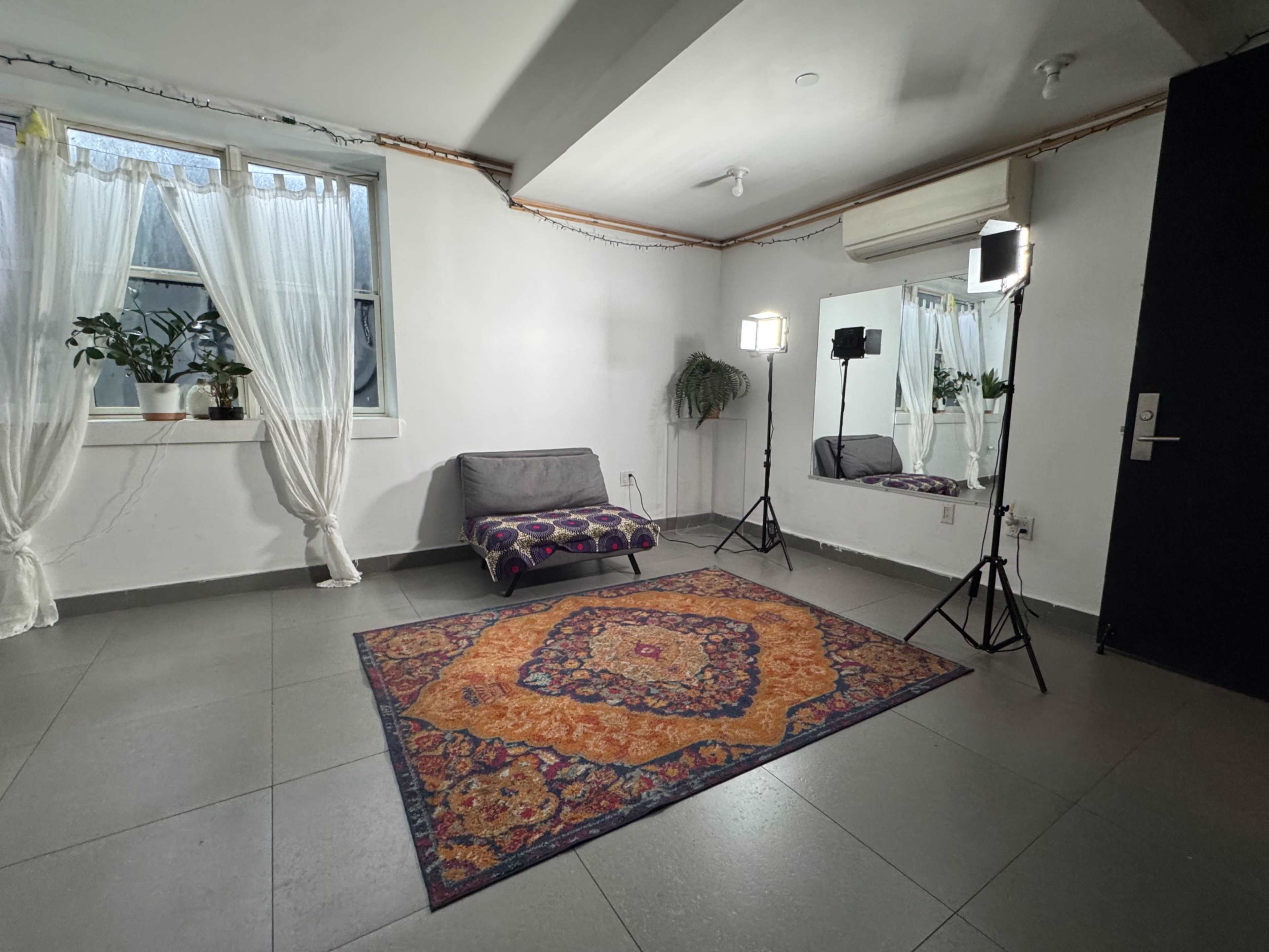 The image shows a sparsely furnished room with a large area rug, a gray sofa, two softbox lights, and windows draped with white curtains.