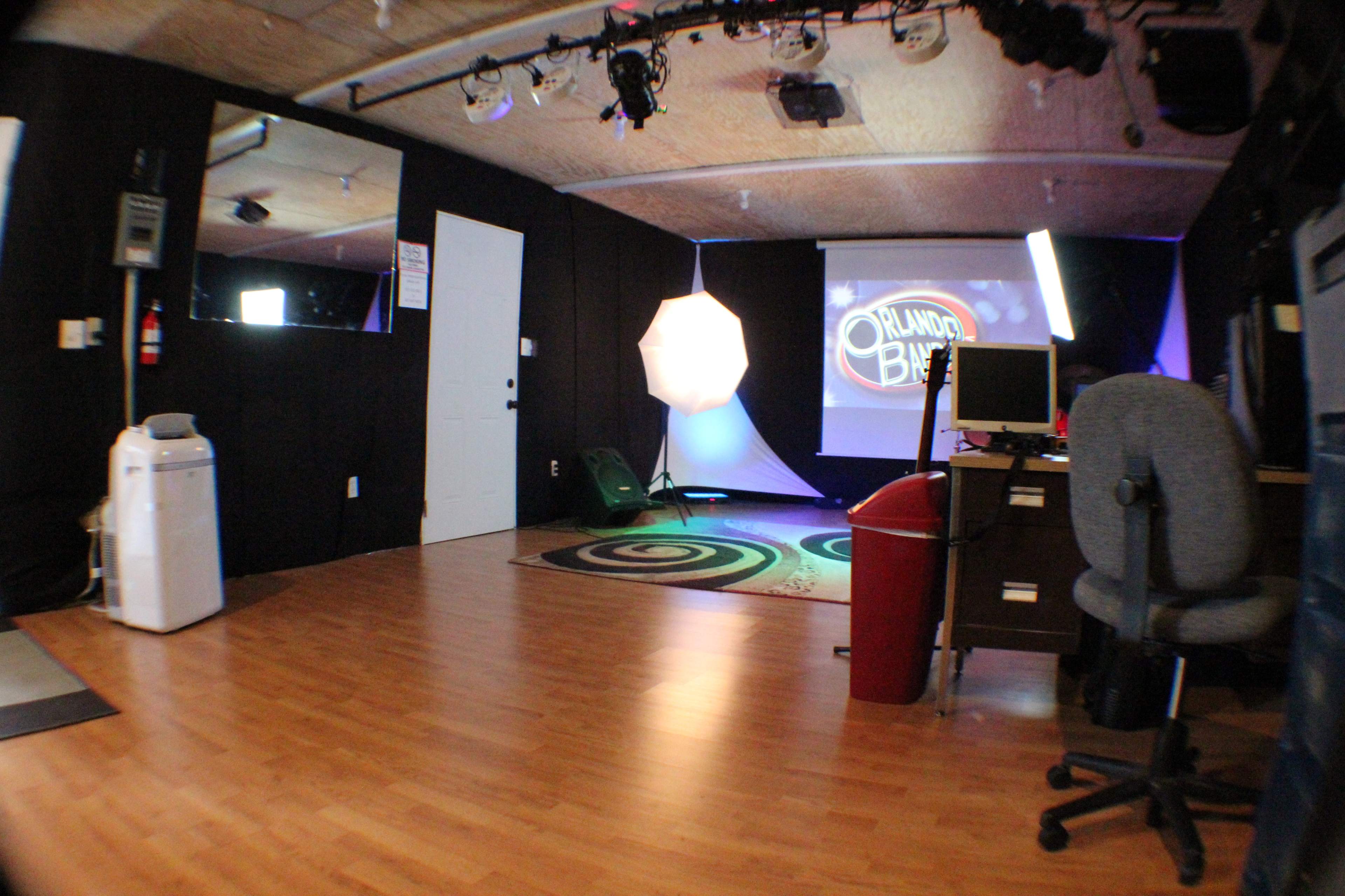 The image shows a small studio space with a wooden floor, a gray office chair, a desk with a computer, and lighting equipment set up in front of a backdrop featuring the words "Project B."