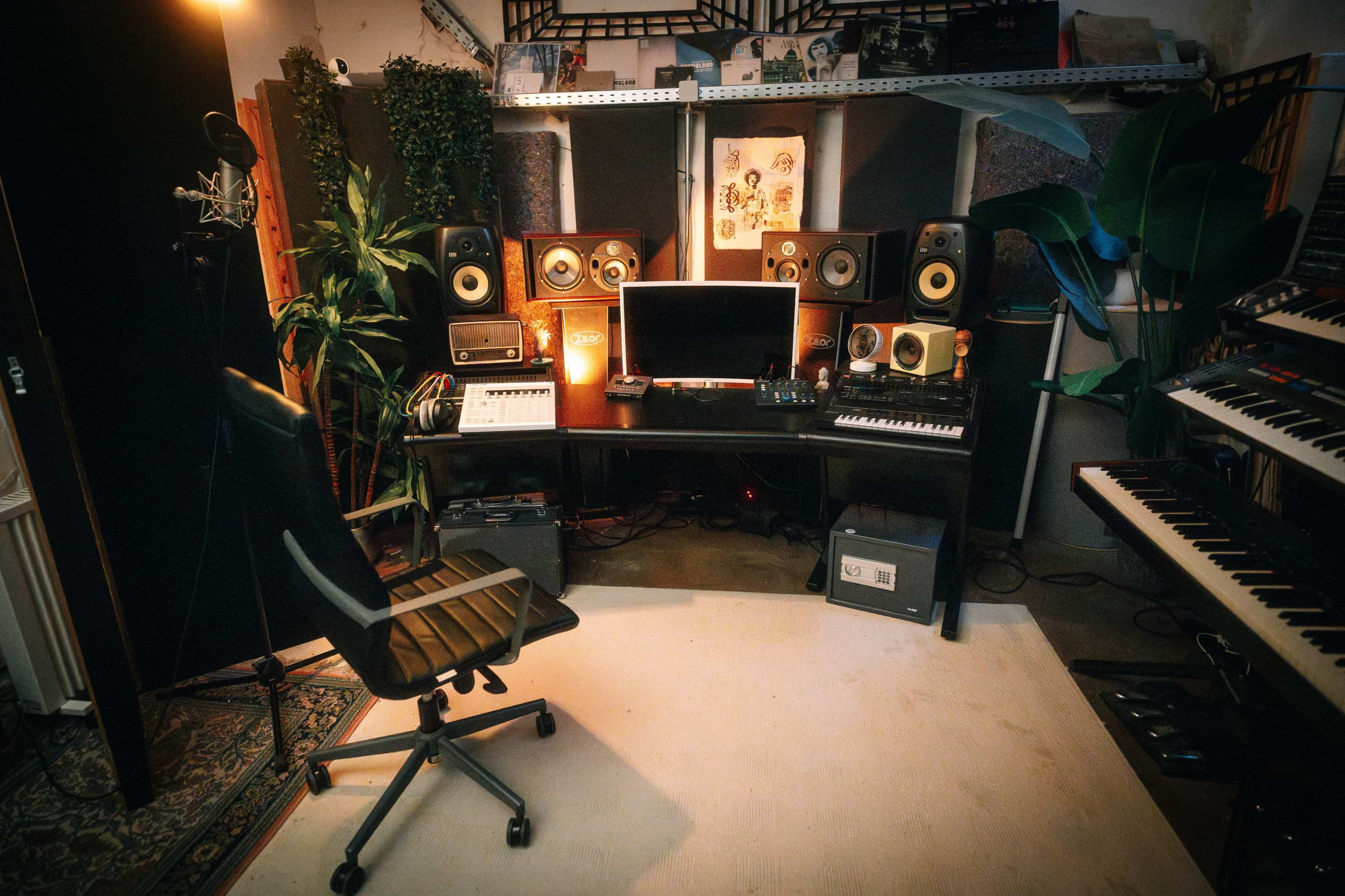 A music production studio features a desk with monitors, a keyboard, and various audio equipment, surrounded by plants and acoustic panels.