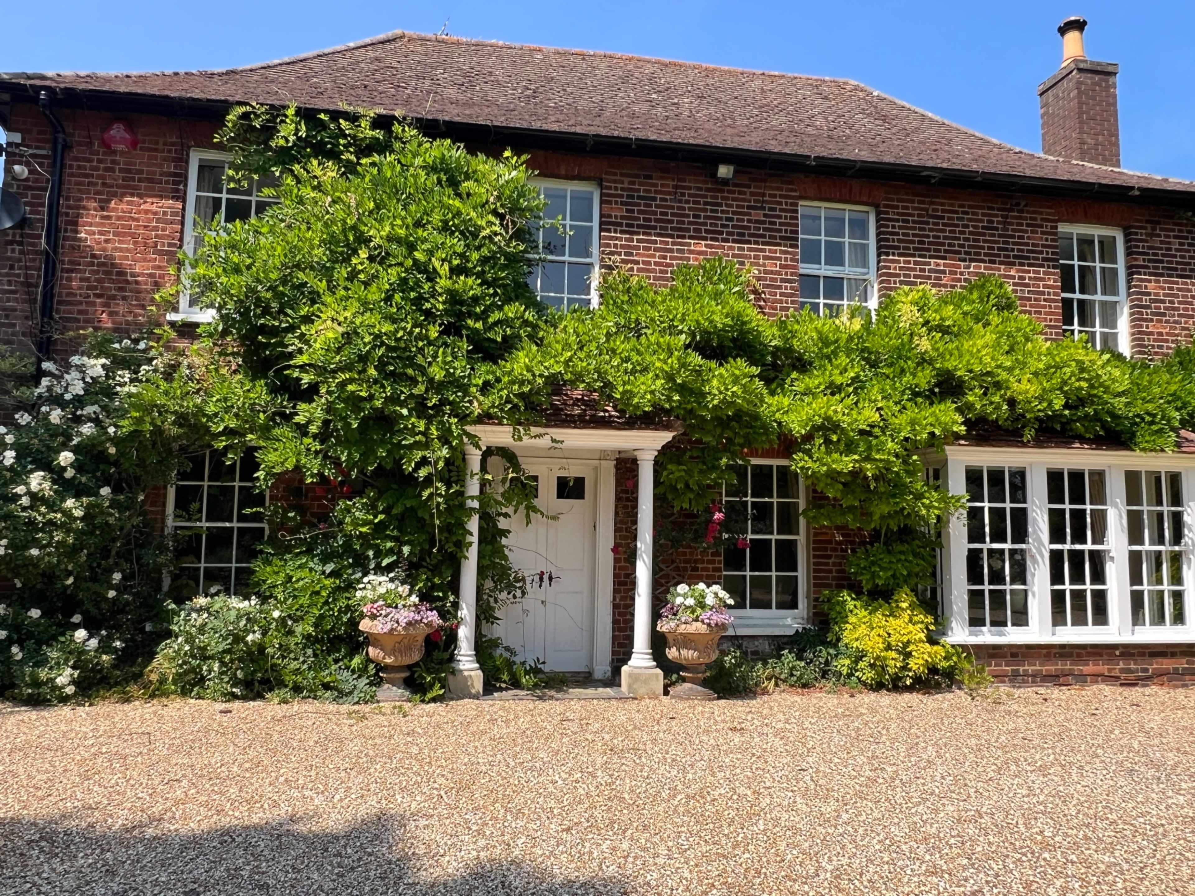 A brick house with lush green vines and shrubs surrounds its entrance, featuring white pillars and large windows.