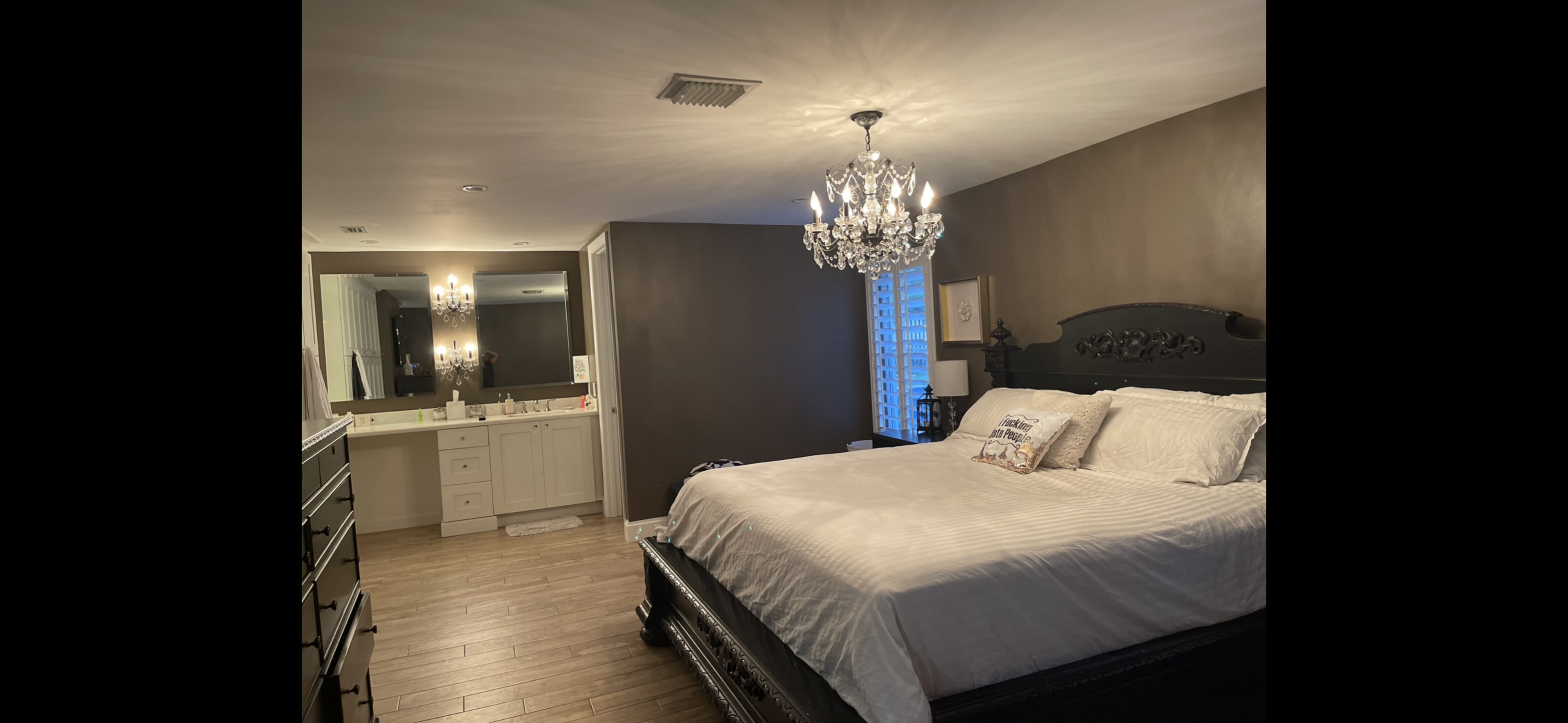 The image shows a spacious bedroom featuring a large bed with a chandelier overhead and a bathroom area visible in the background.