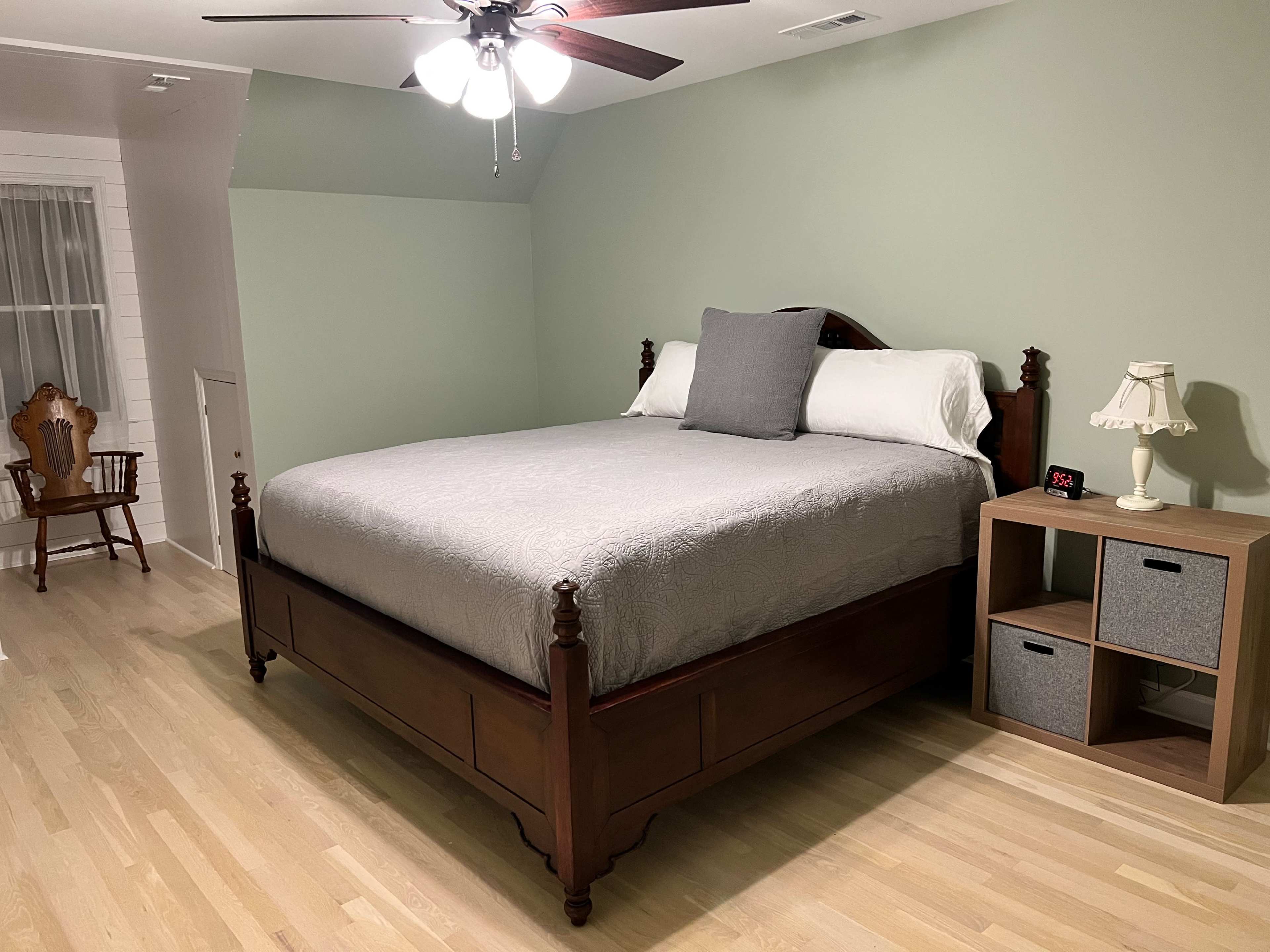 The image shows a bedroom with a wooden bed, a gray quilt, a bedside lamp, and a small nightstand with storage boxes.