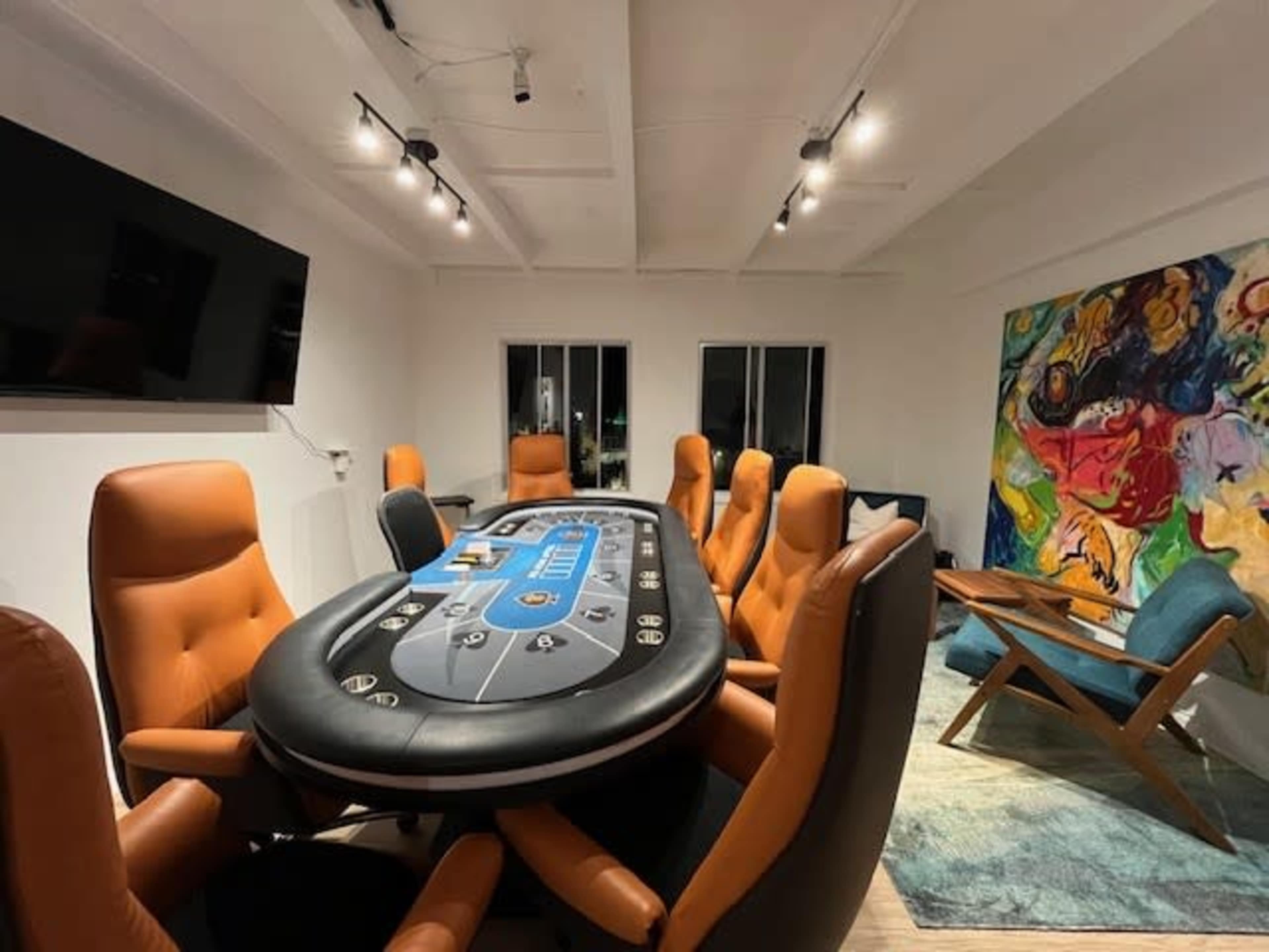A poker table with eight brown leather chairs is set up in a brightly lit room featuring a large abstract painting and a blue upholstered chair.