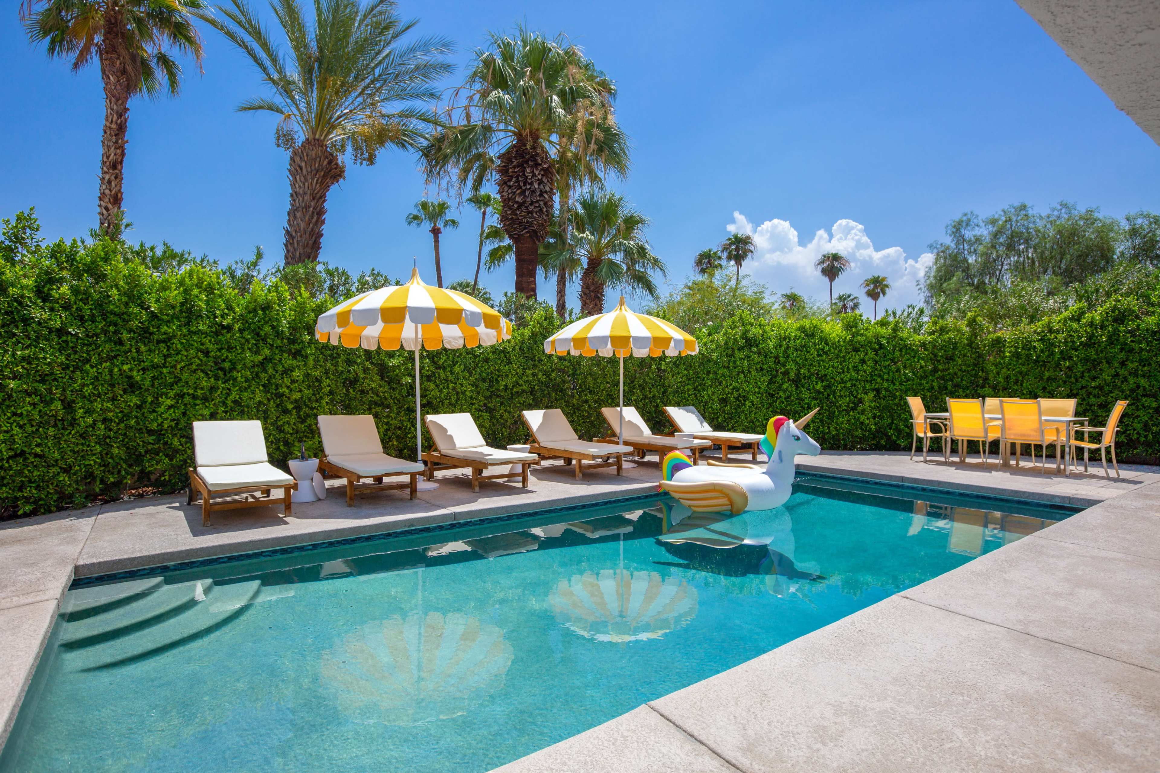 A swimming pool surrounded by lounge chairs, yellow and white umbrellas, and a large inflatable unicorn in a tropical setting.