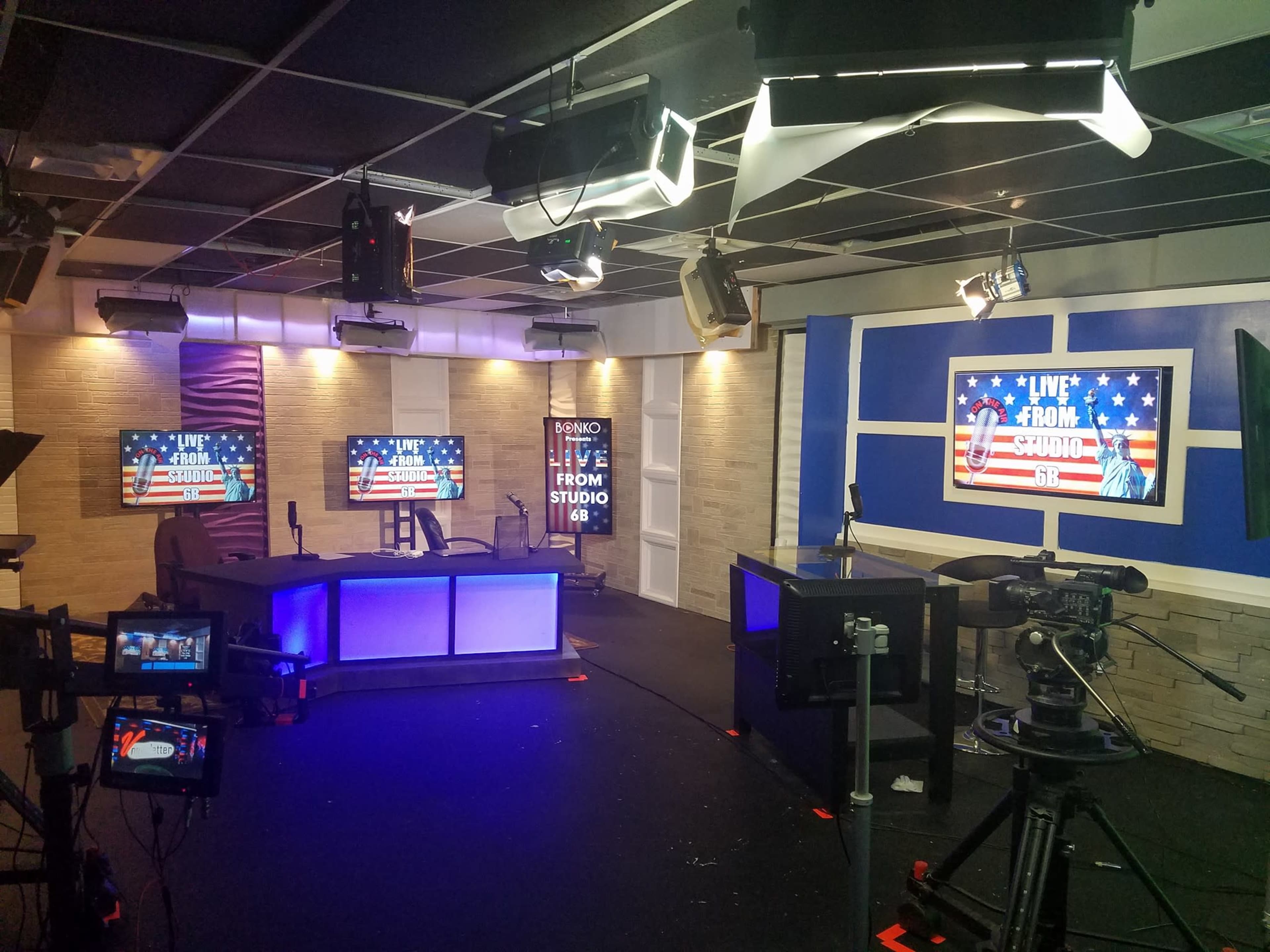 The image shows a television studio setup with multiple cameras, a central desk, and screens displaying "LIVE FROM STUDIO 16" against a patriotic background.
