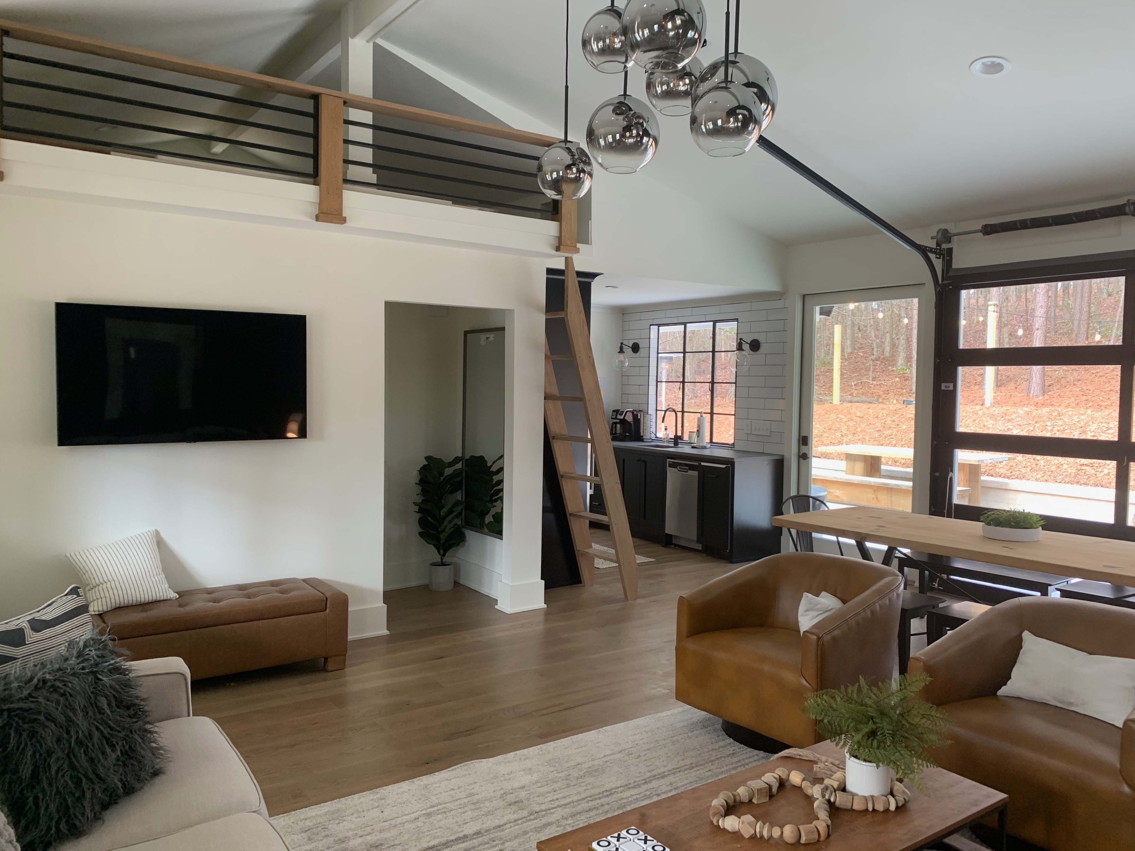 A modern living space with a sectional sofa, a wall-mounted television, a dining area, and a kitchen, all featuring large windows that overlook a wooded outdoor space.