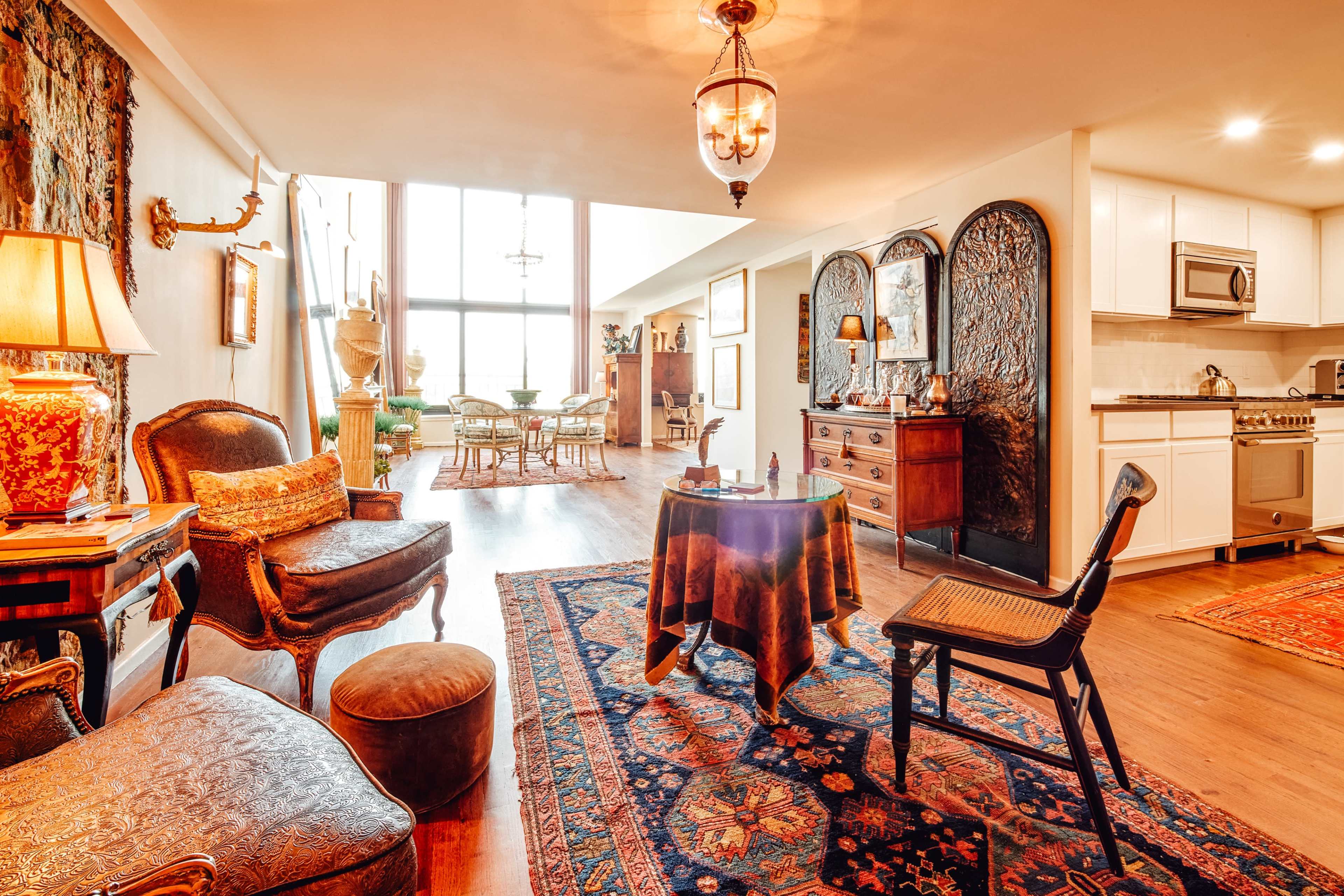 The image shows a spacious living area with elegant furniture, a patterned rug, and large windows allowing natural light to enter.