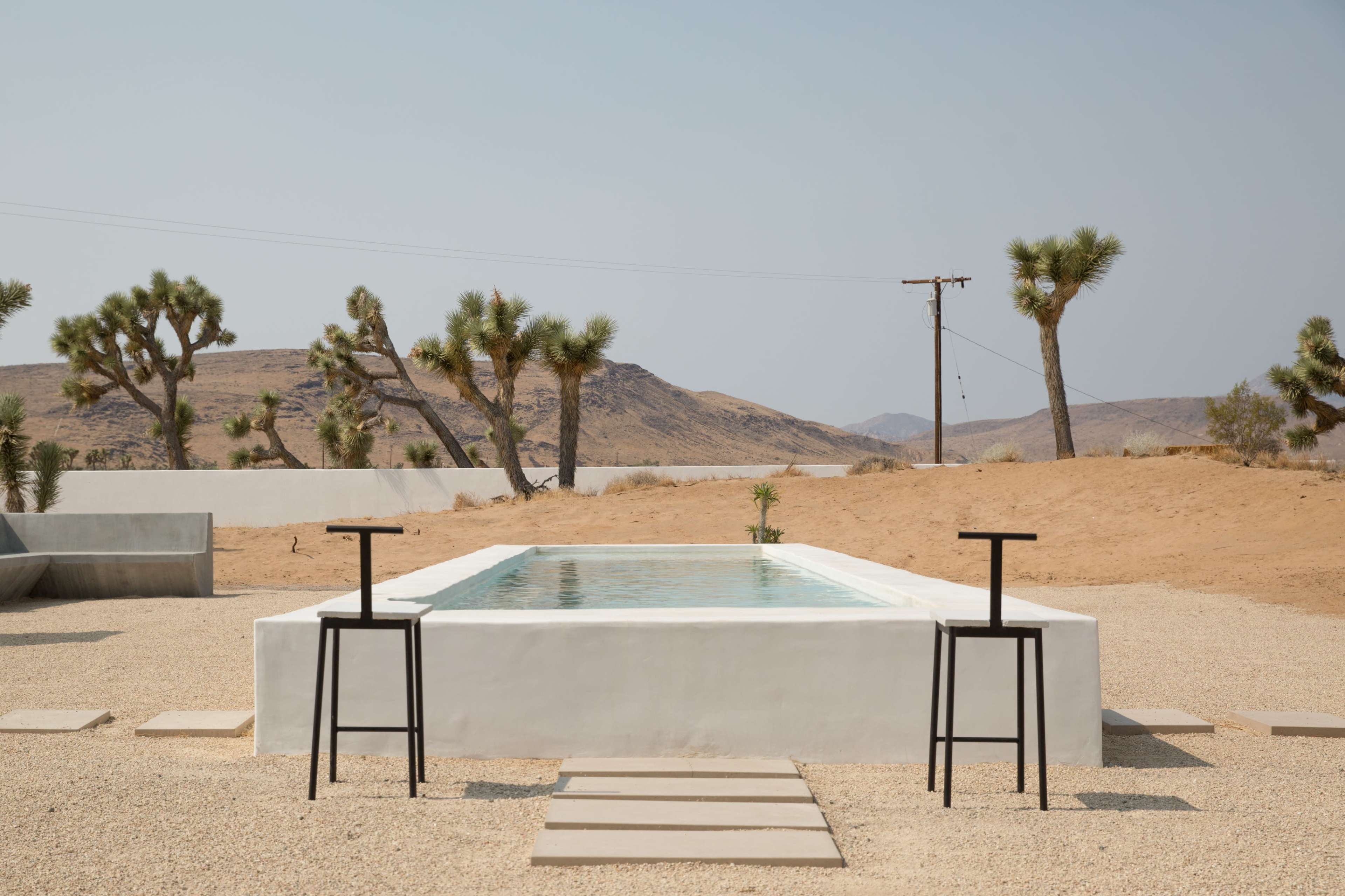 A rectangular pool sits in a minimalist desert setting, flanked by two black chairs and surrounded by rocky terrain and sparse vegetation.
