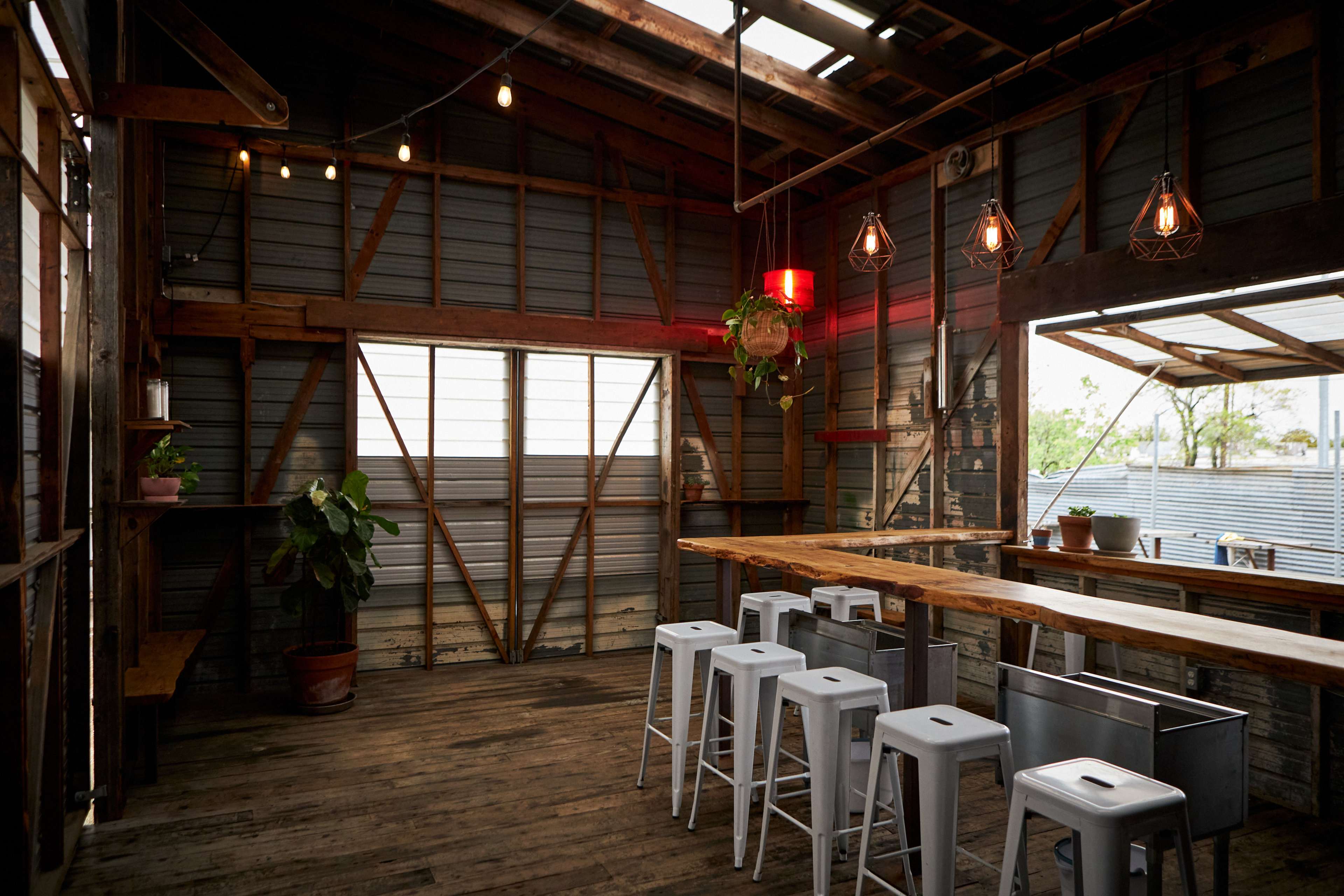 An interior of a rustic space with wooden beams, a long table, and several metal stools, illuminated by pendant lights.