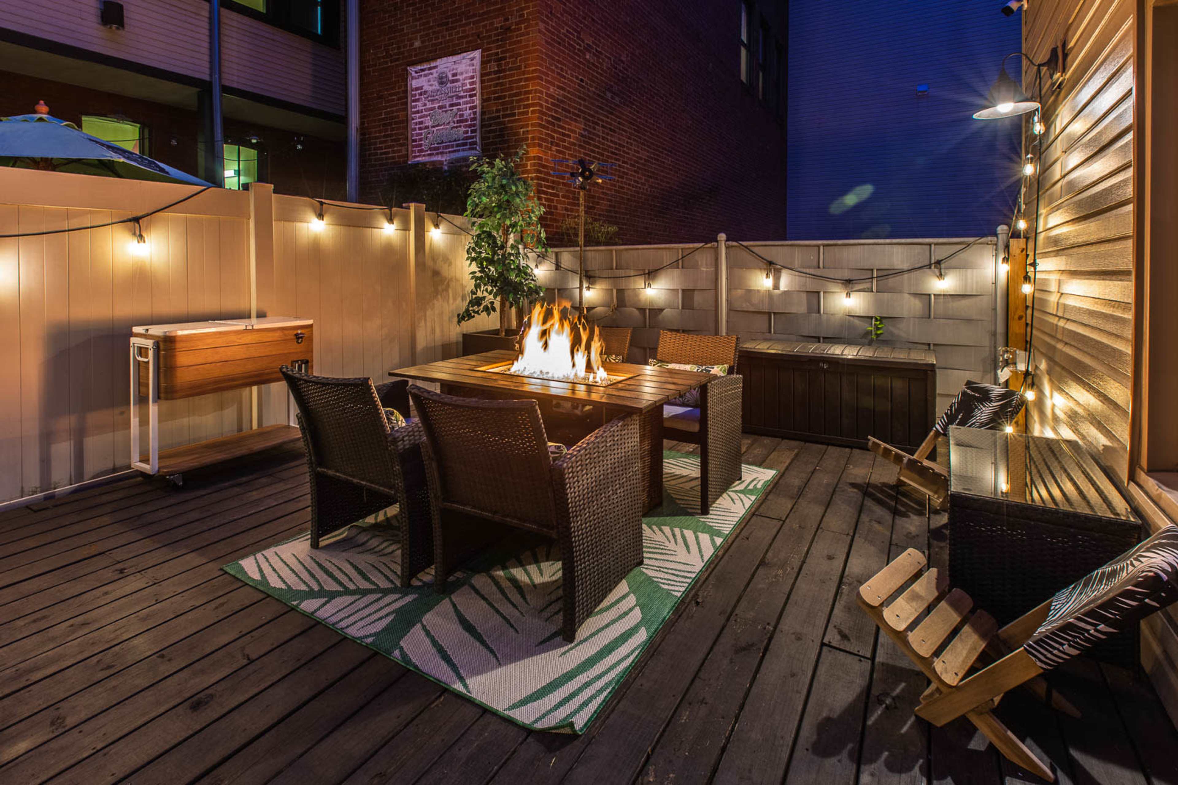 A cozy outdoor patio features a fire table surrounded by four wicker chairs, illuminated by string lights and set against a wooden deck.