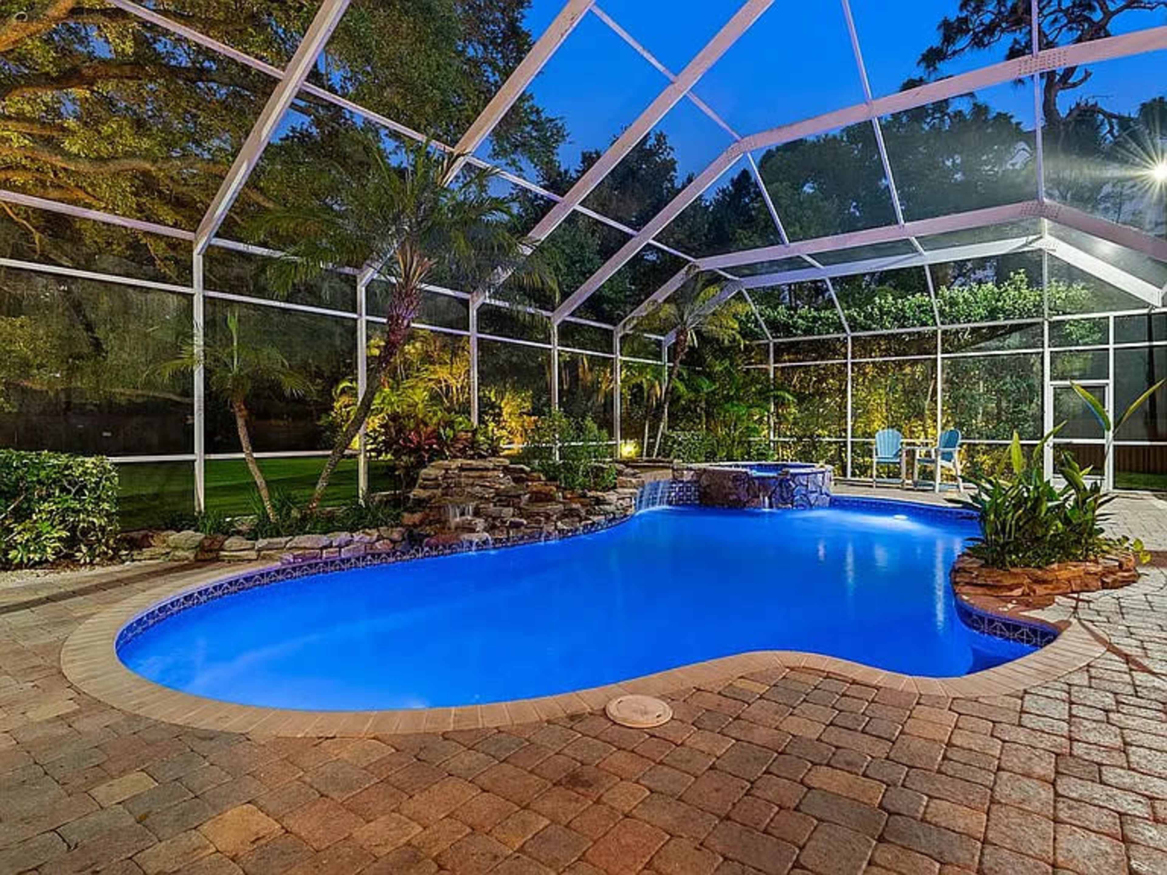 A swimming pool with a rock waterfall is enclosed in a screened patio surrounded by lush greenery.