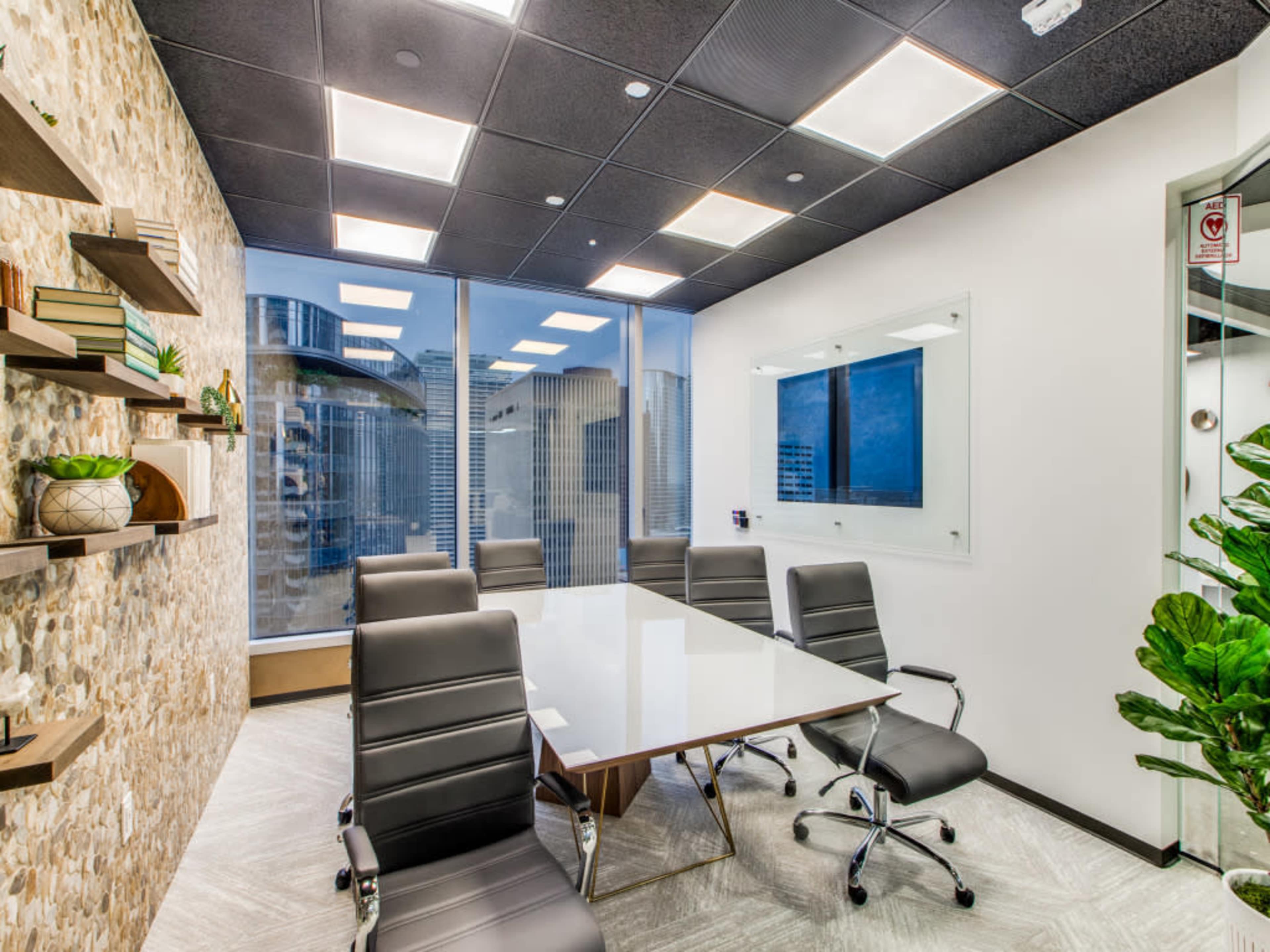 A modern conference room with a glass wall, a large white table, and black leather chairs arranged around it, overlooking a city skyline.