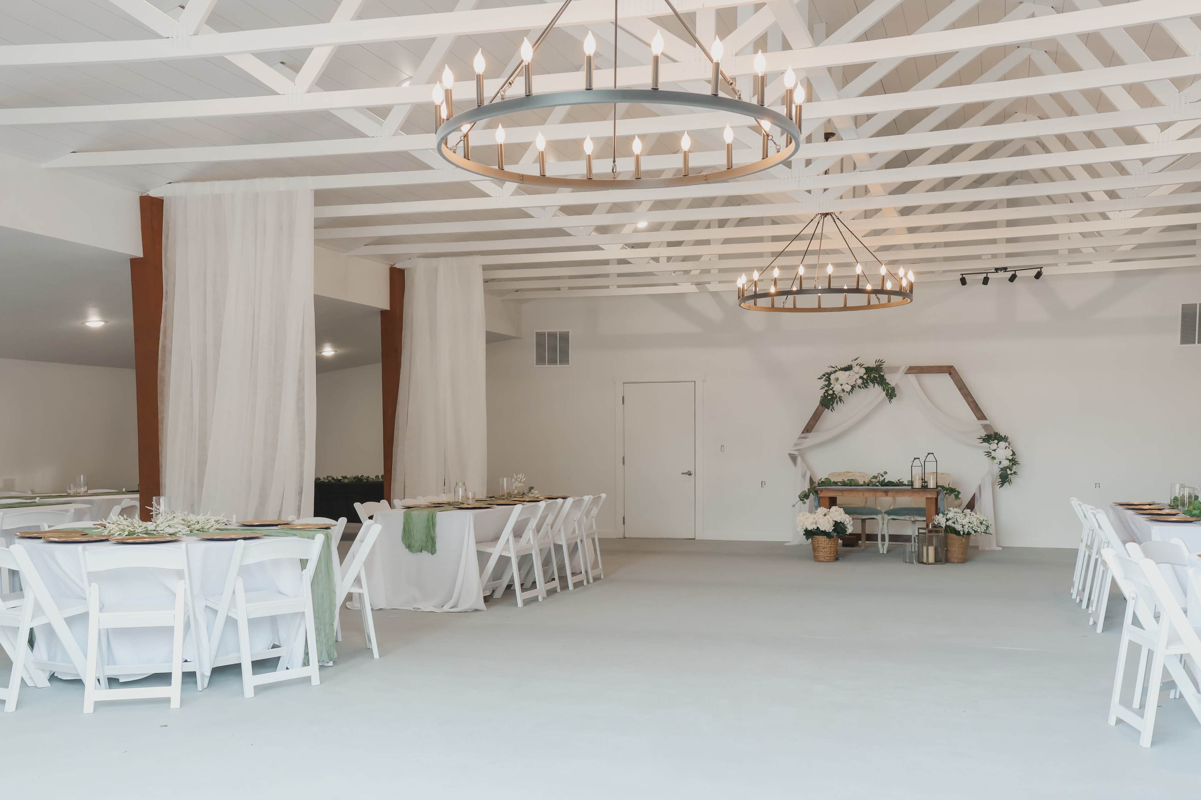 The image depicts a spacious and well-lit event space with white tables and chairs arranged around the room, and a decorated centerpiece table against a plain backdrop.