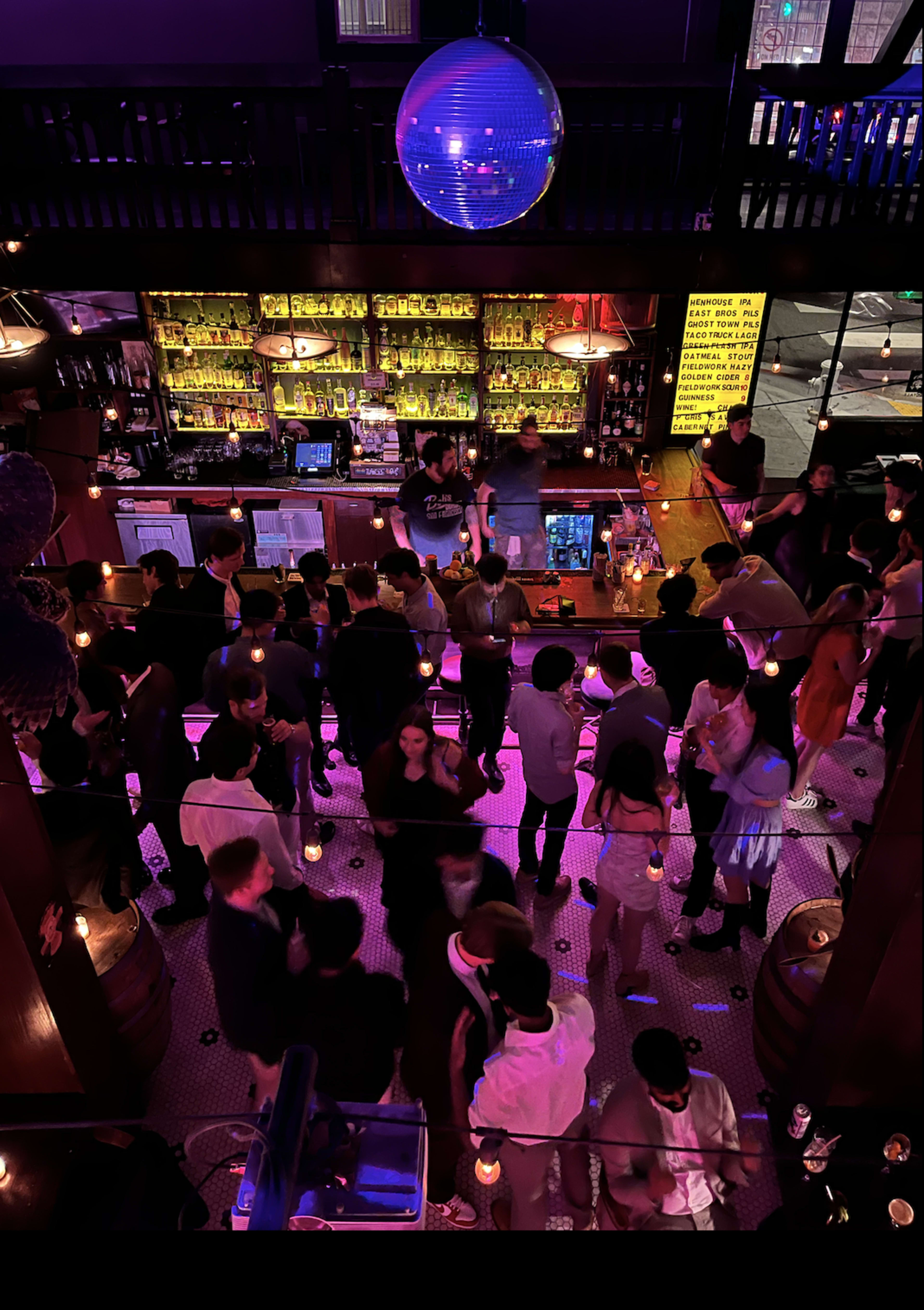A busy bar scene with patrons socializing and a bartender preparing drinks under a large disco ball.