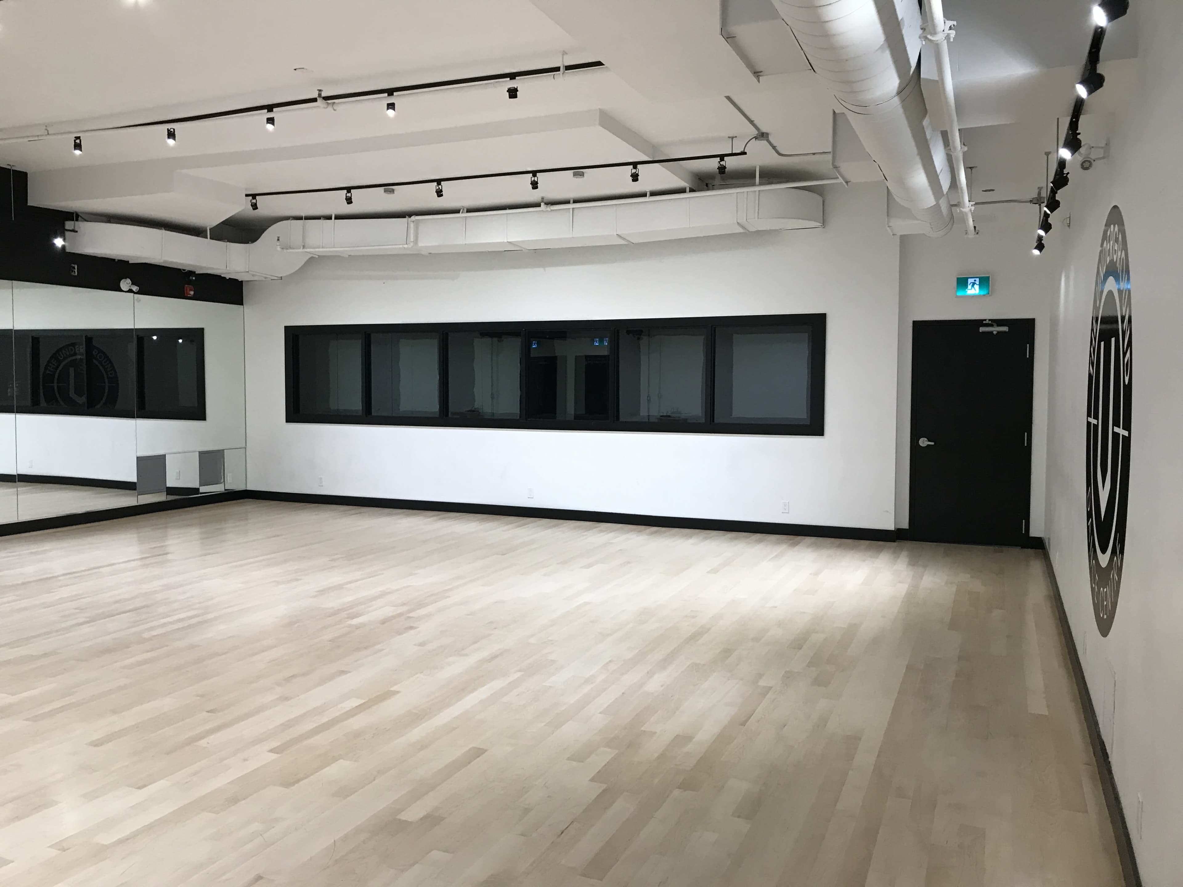 The image shows a spacious dance studio with wooden flooring, large windows, and a mirror along one wall.