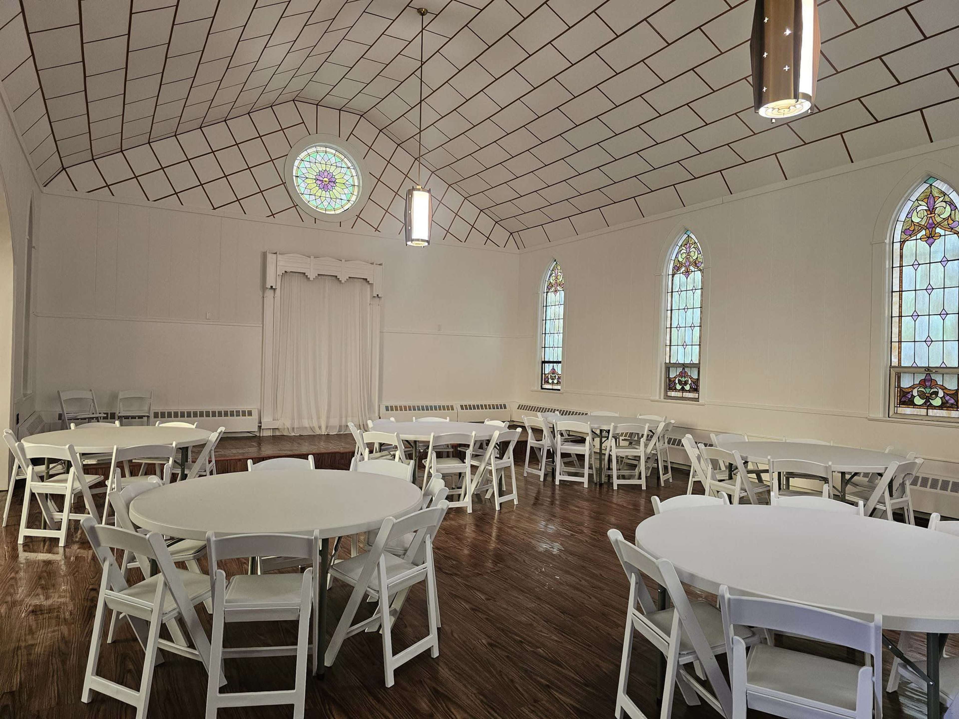 A large, well-lit room with several round tables surrounded by white chairs, featuring stained glass windows and a simple stage area at one end.