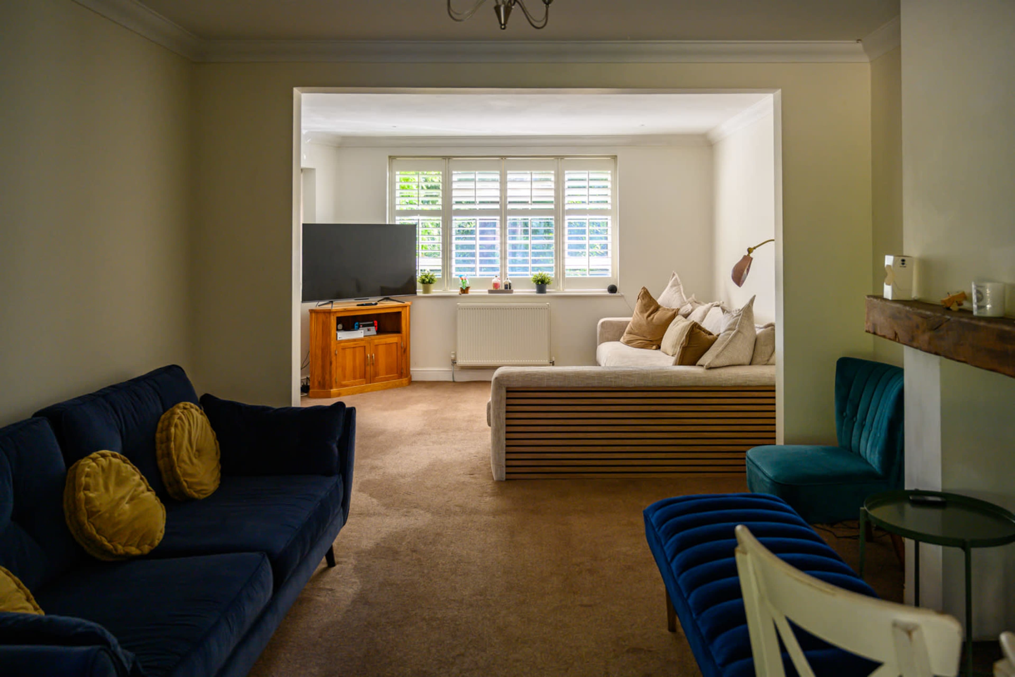 A living room features a blue sofa, a green chair, a cream couch, and a television cabinet, all arranged around a light brown carpet.