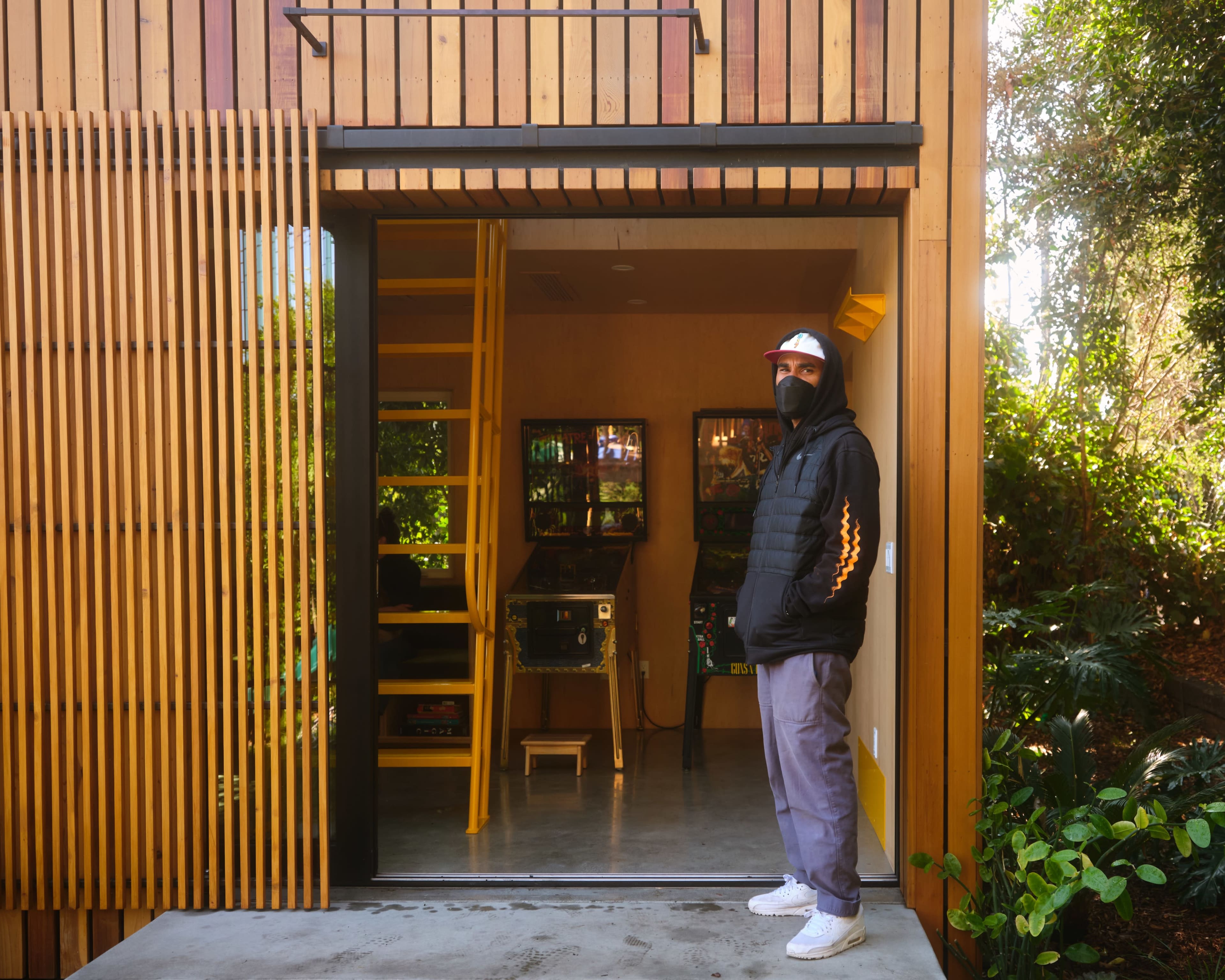 A person wearing a black coat and mask stands beside an entrance to a wooden building with pinball machines visible inside.