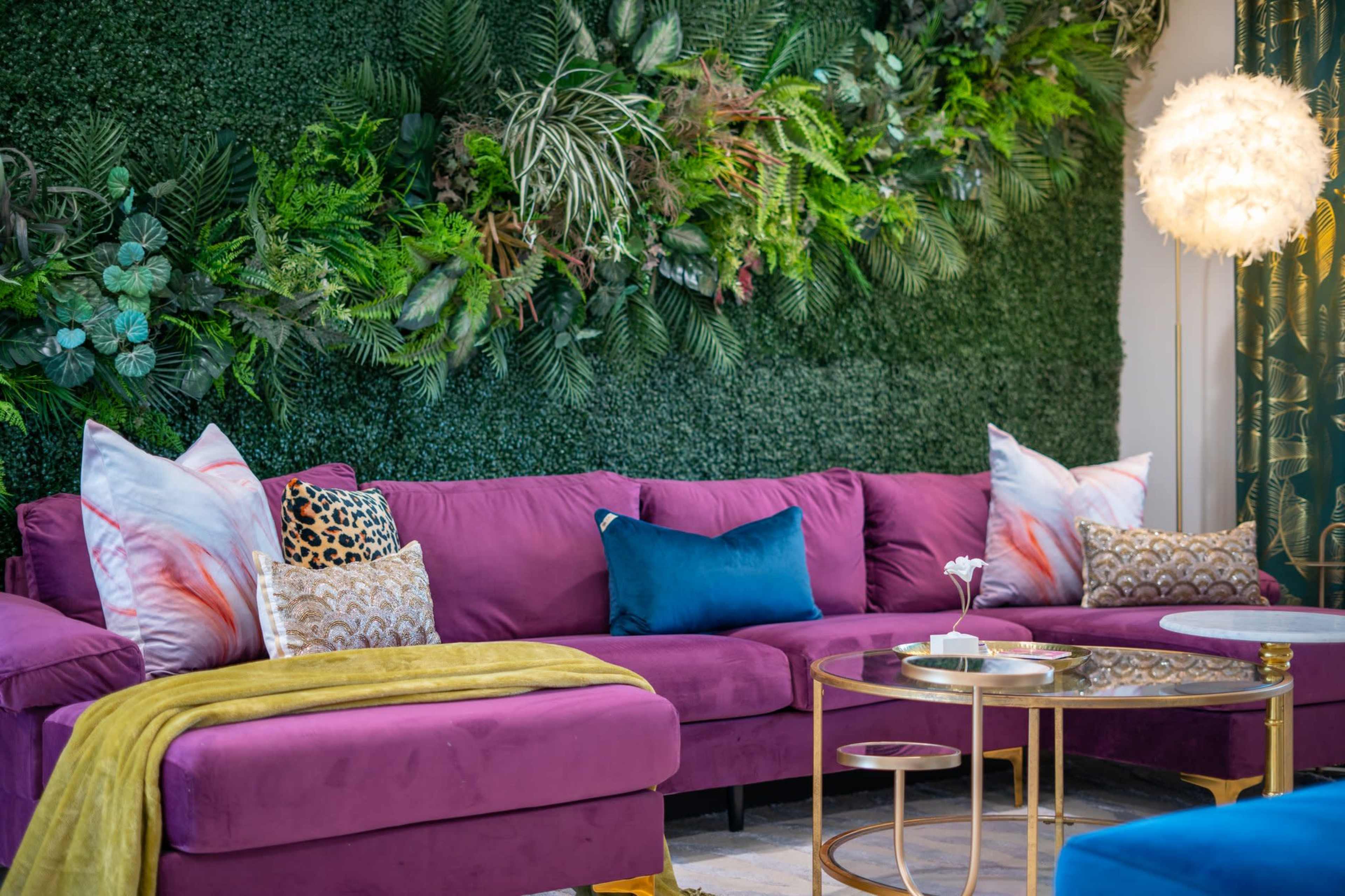 A modern living room features a purple sectional sofa, decorative pillows, a green vertical garden backdrop, and a round gold coffee table.