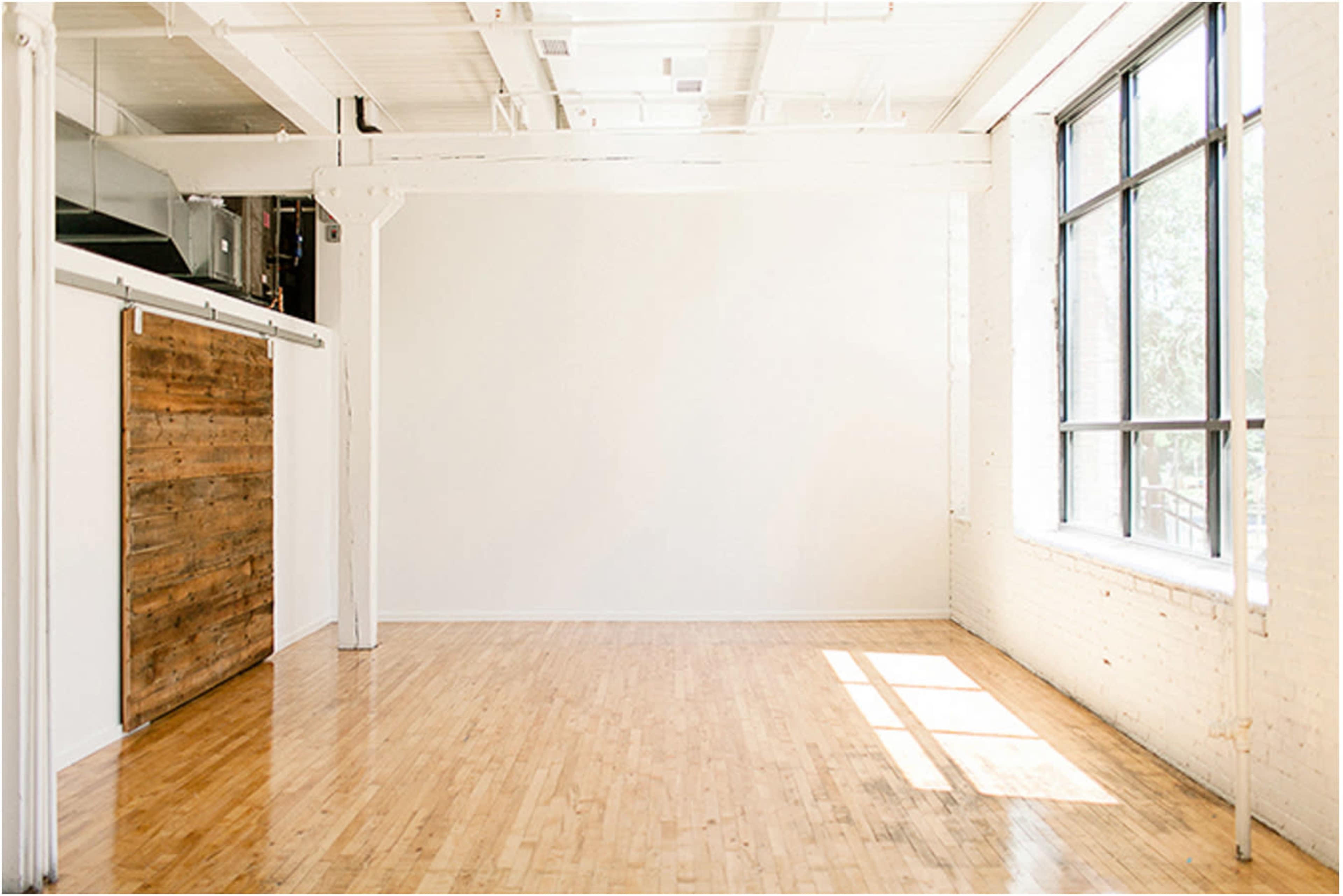 The image shows an empty room with wooden flooring, a large window letting in natural light, and a sliding barn door against one wall.