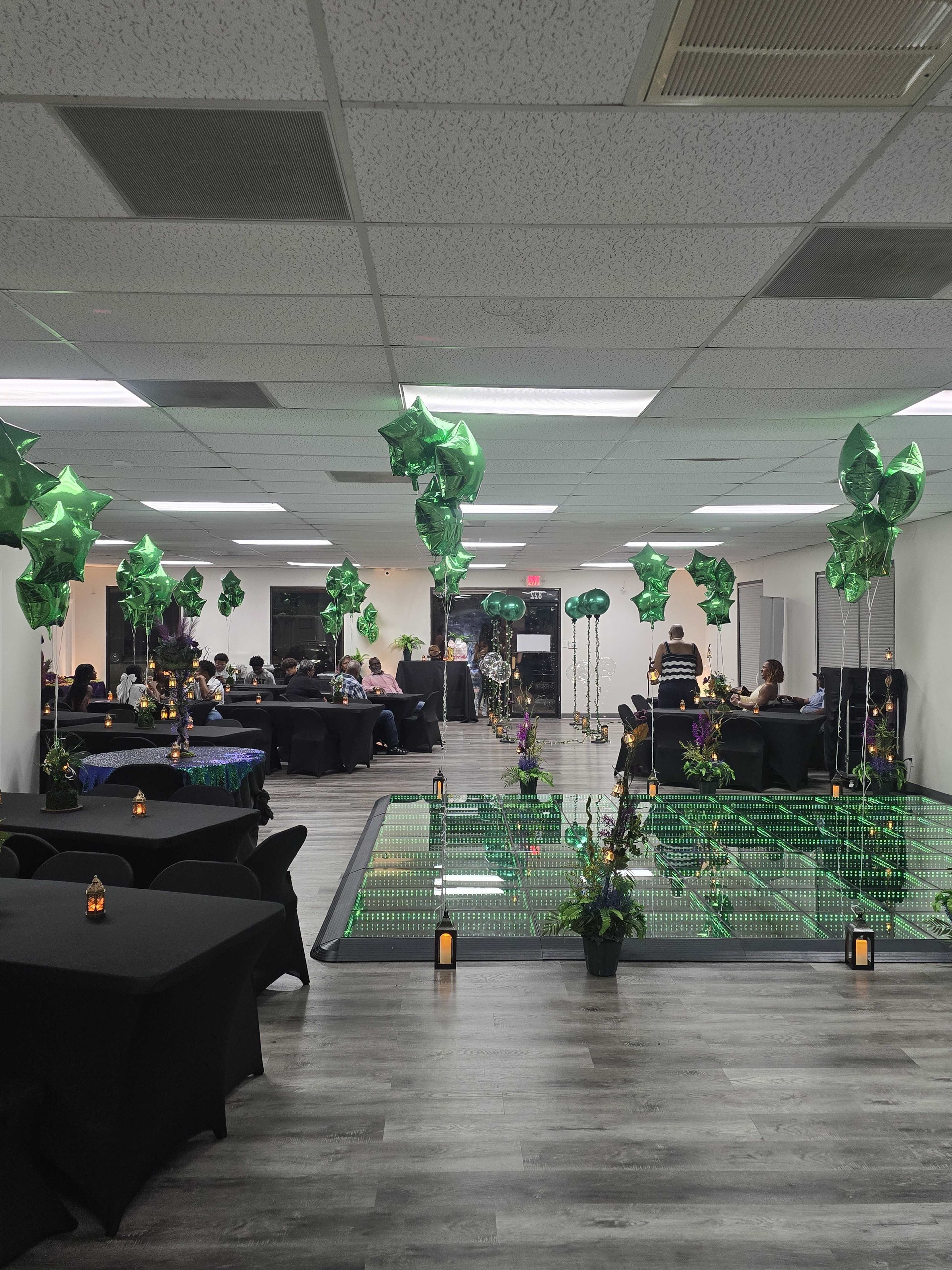 The image shows an indoor event space decorated with green star-shaped balloons and tables arranged around a central area with plants and candles.