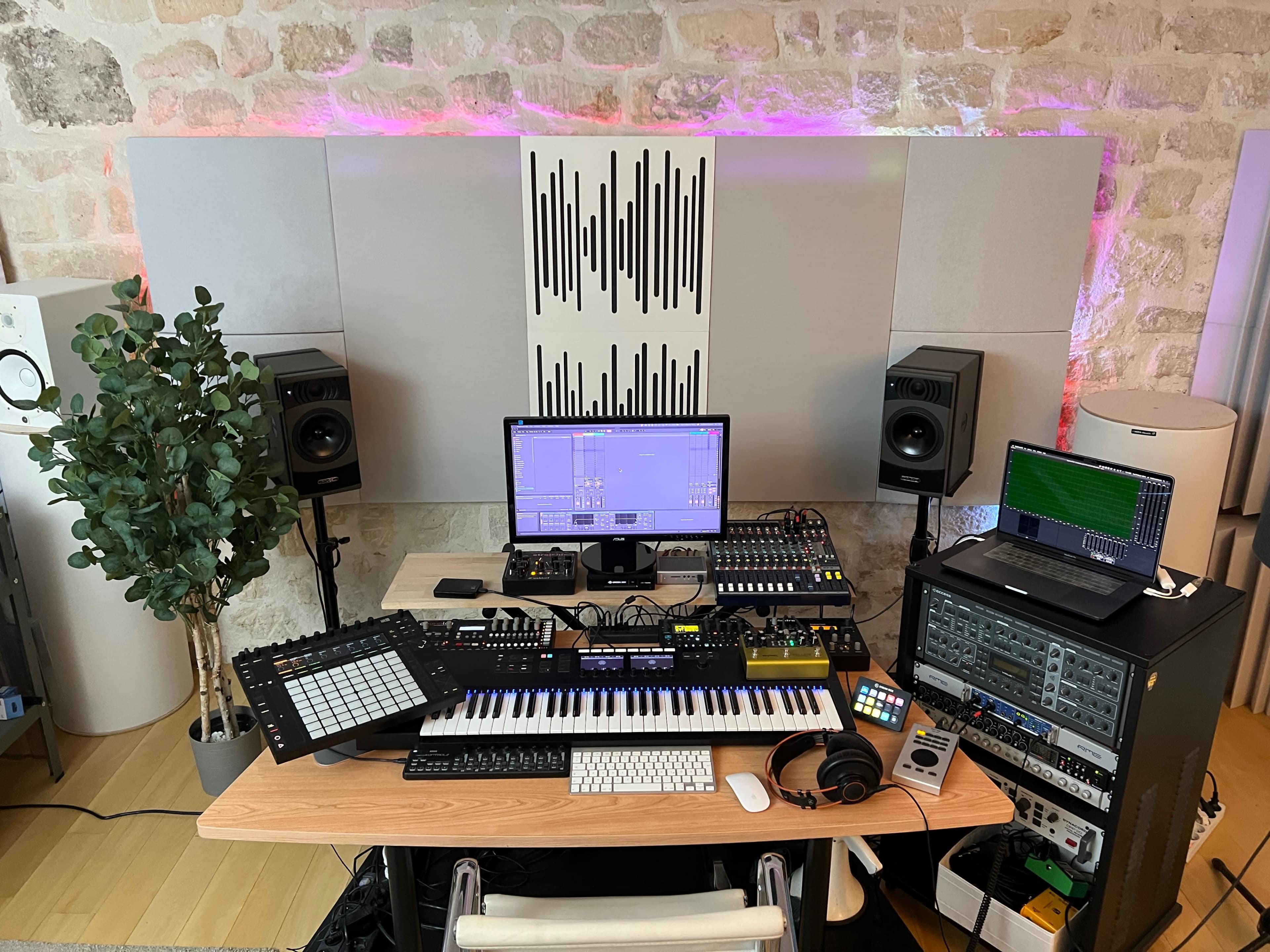 A digital music production setup featuring a keyboard, monitors, computer, audio interface, and various controllers, set against a stone wall.