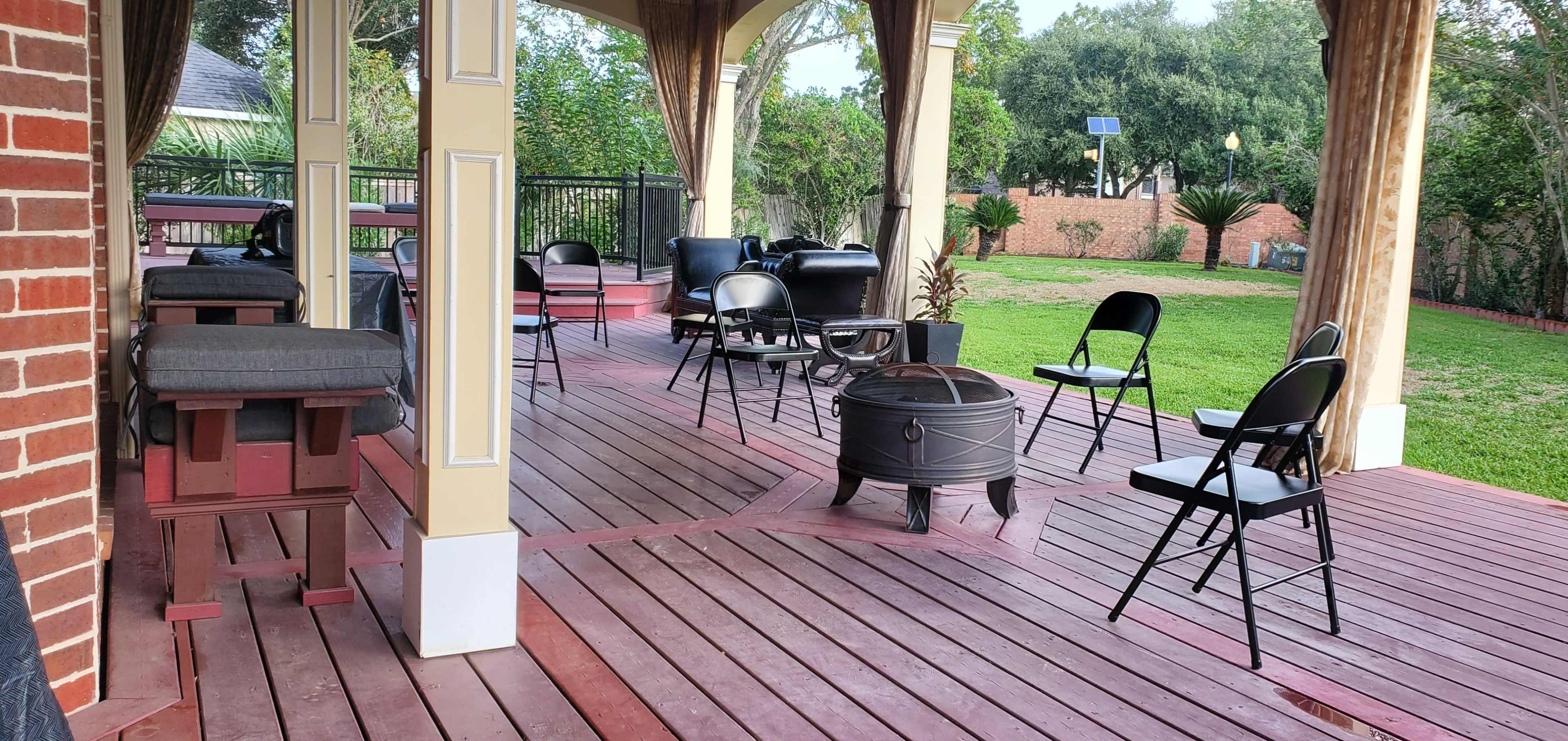 A covered wooden patio with several black folding chairs arranged around a fire pit and potted plants in the background.