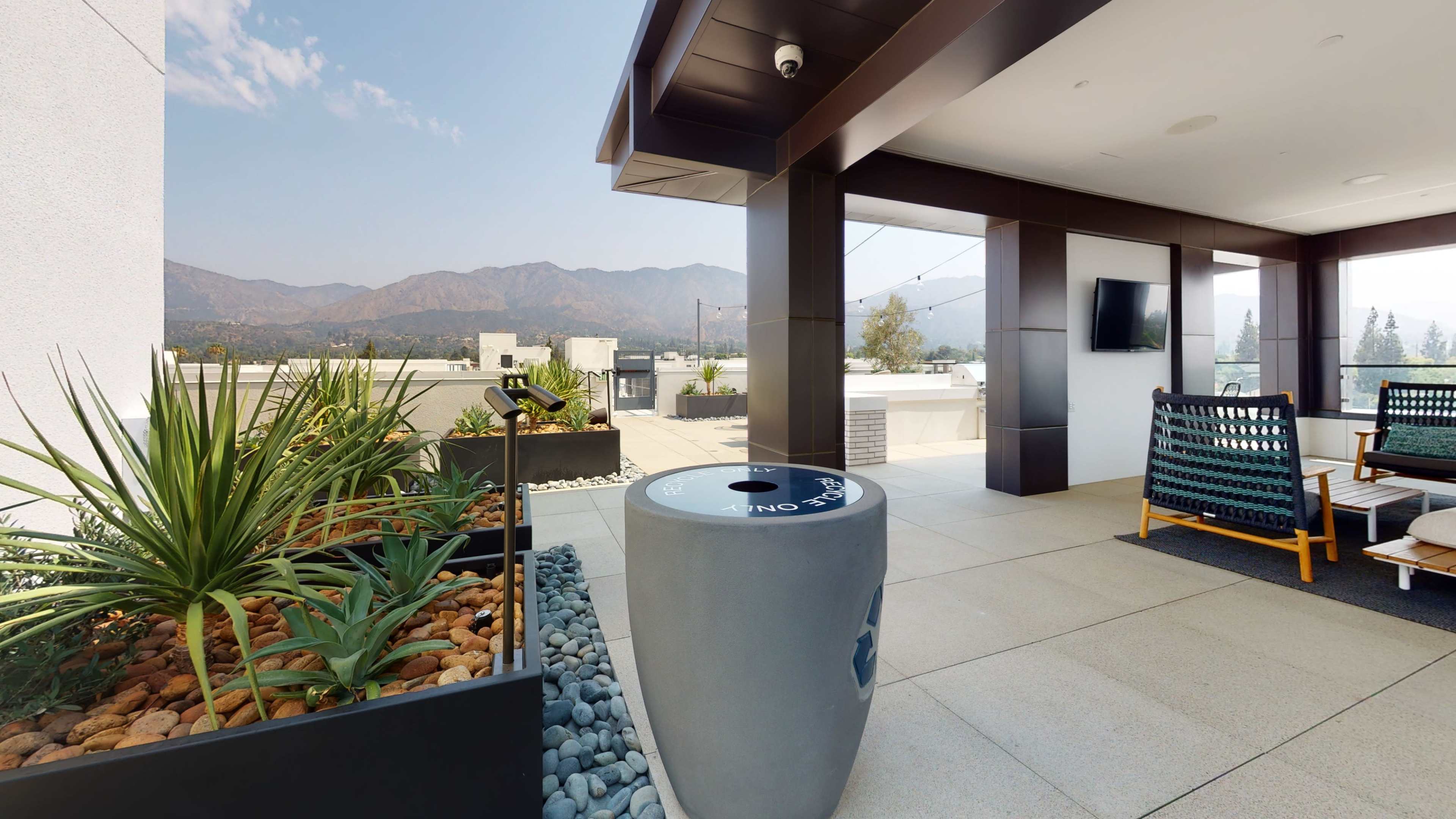 A rooftop terrace features seating areas, a modern trash receptacle, and desert plants with mountains visible in the background.