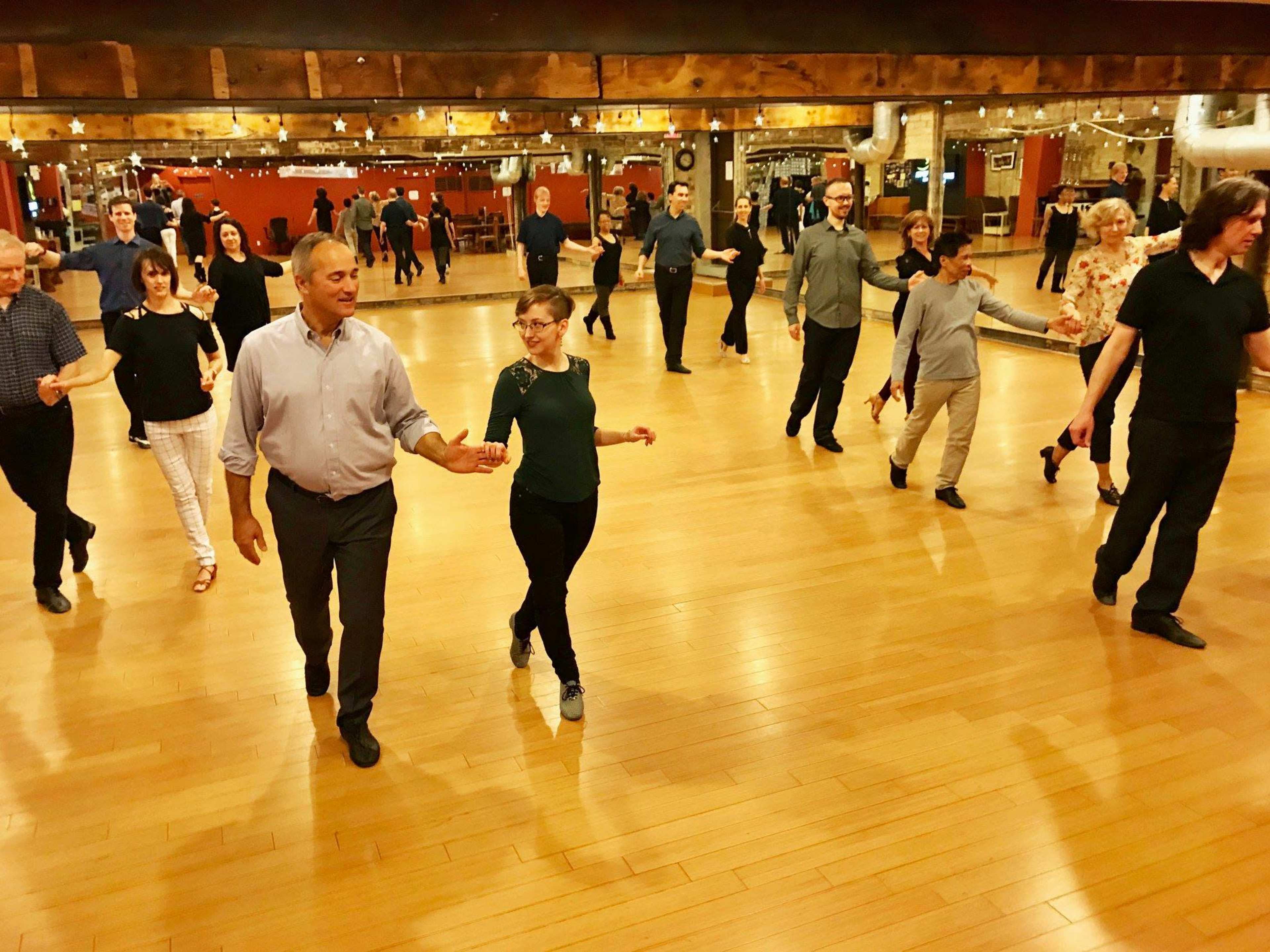 A group of people is dancing in pairs on a wooden floor in a well-lit dance studio.