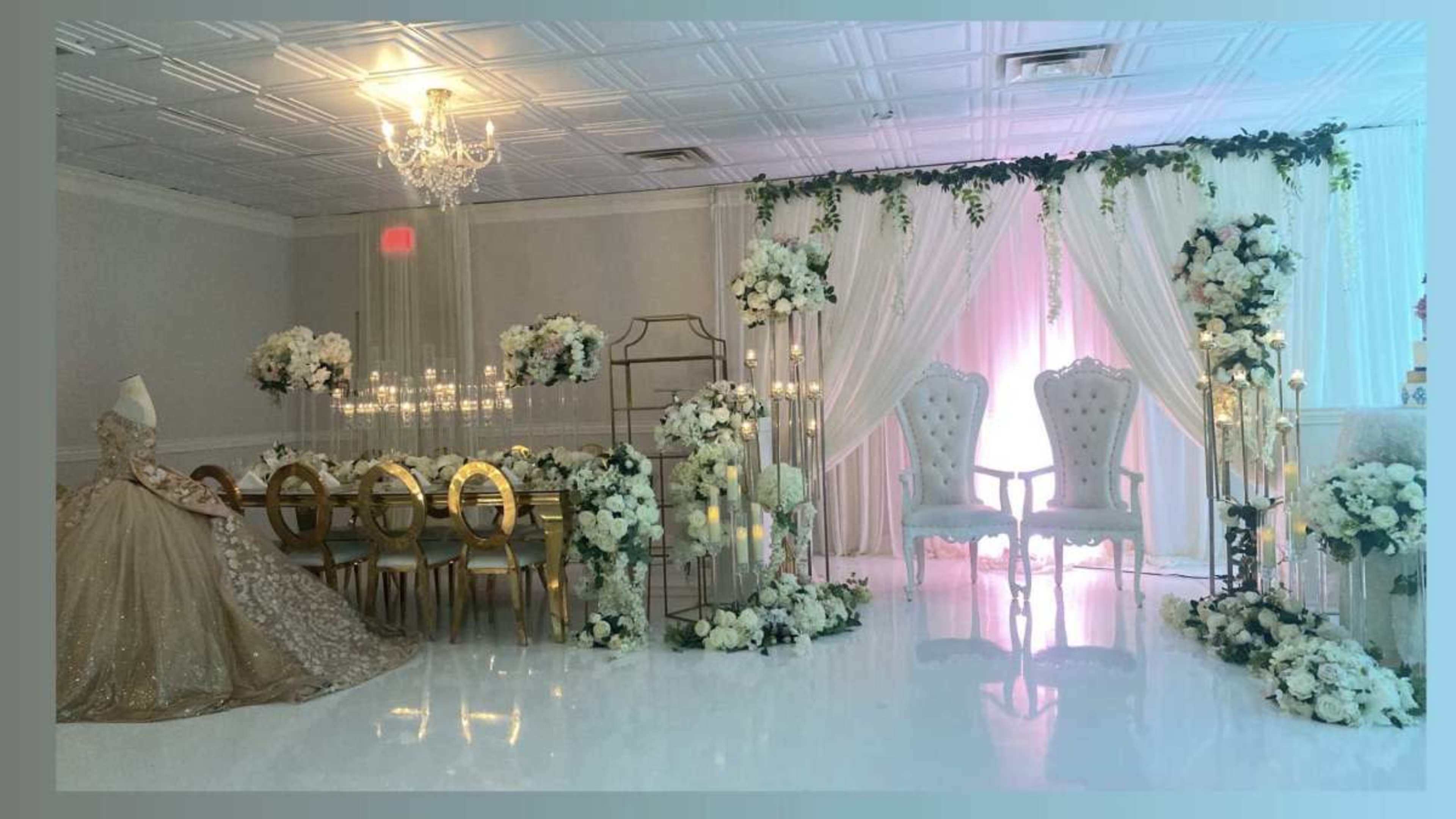 The image shows a decorated indoor space for an event, featuring a long table surrounded by elegant chairs, floral arrangements, and a backdrop with white drapes and golden accents.