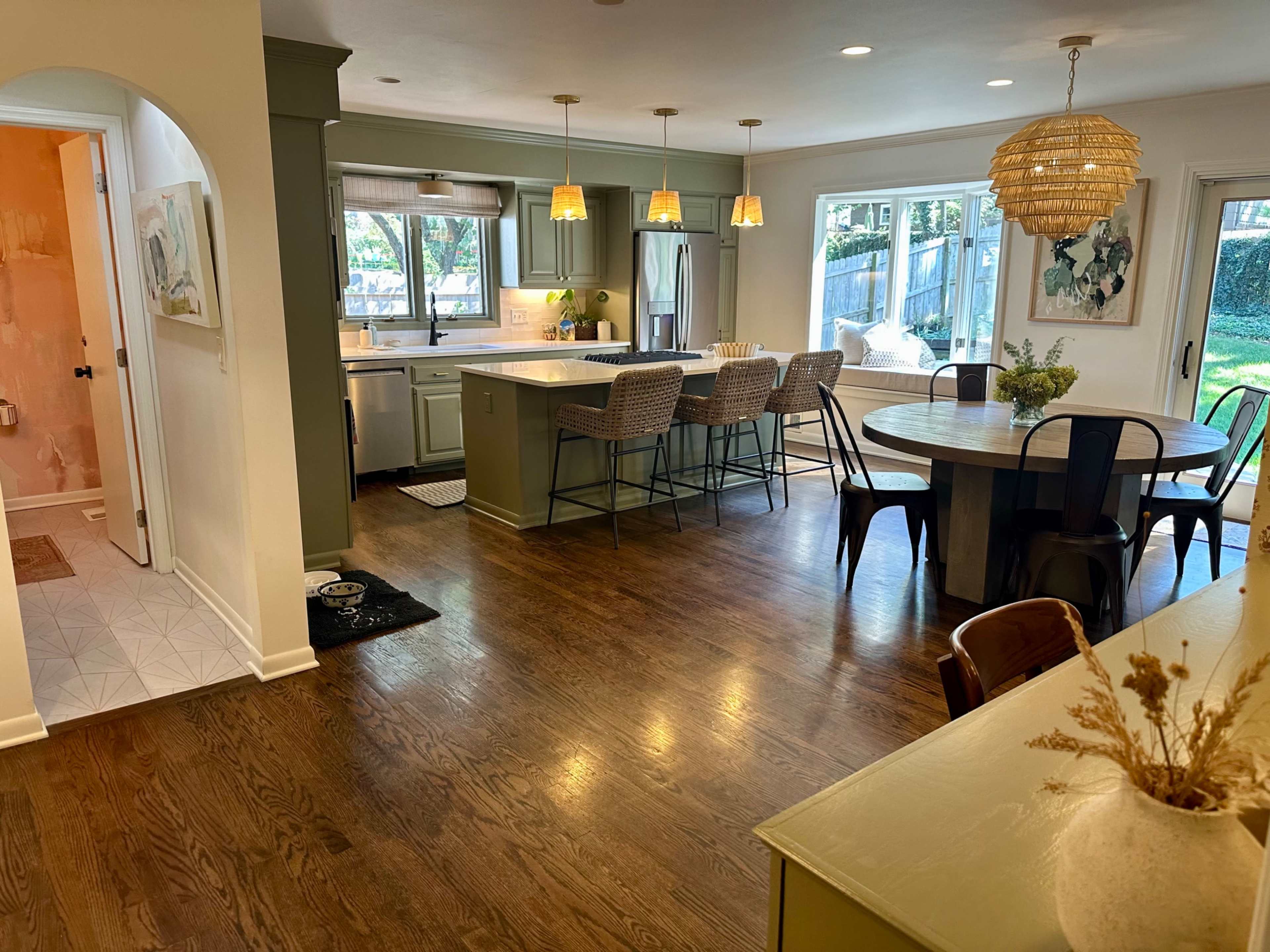 The image shows a modern kitchen and dining area featuring a central island with bar stools, a round dining table, and large windows that allow natural light to fill the space.