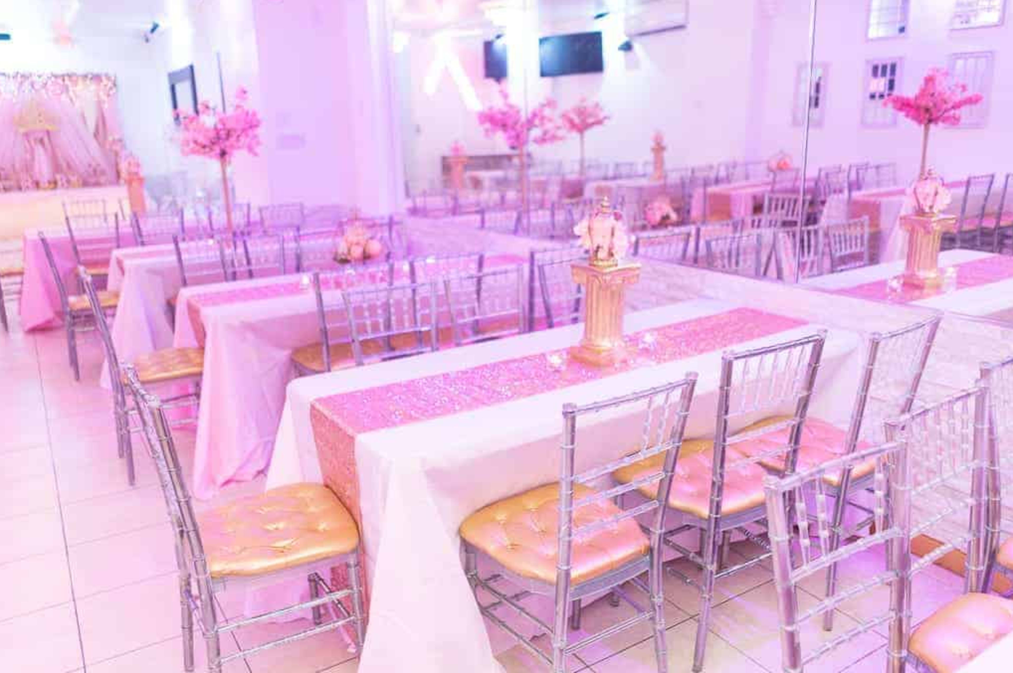 The image shows a banquet hall decorated with pink-themed table settings, featuring white tables with pink runners and silver chairs arranged in neat rows.