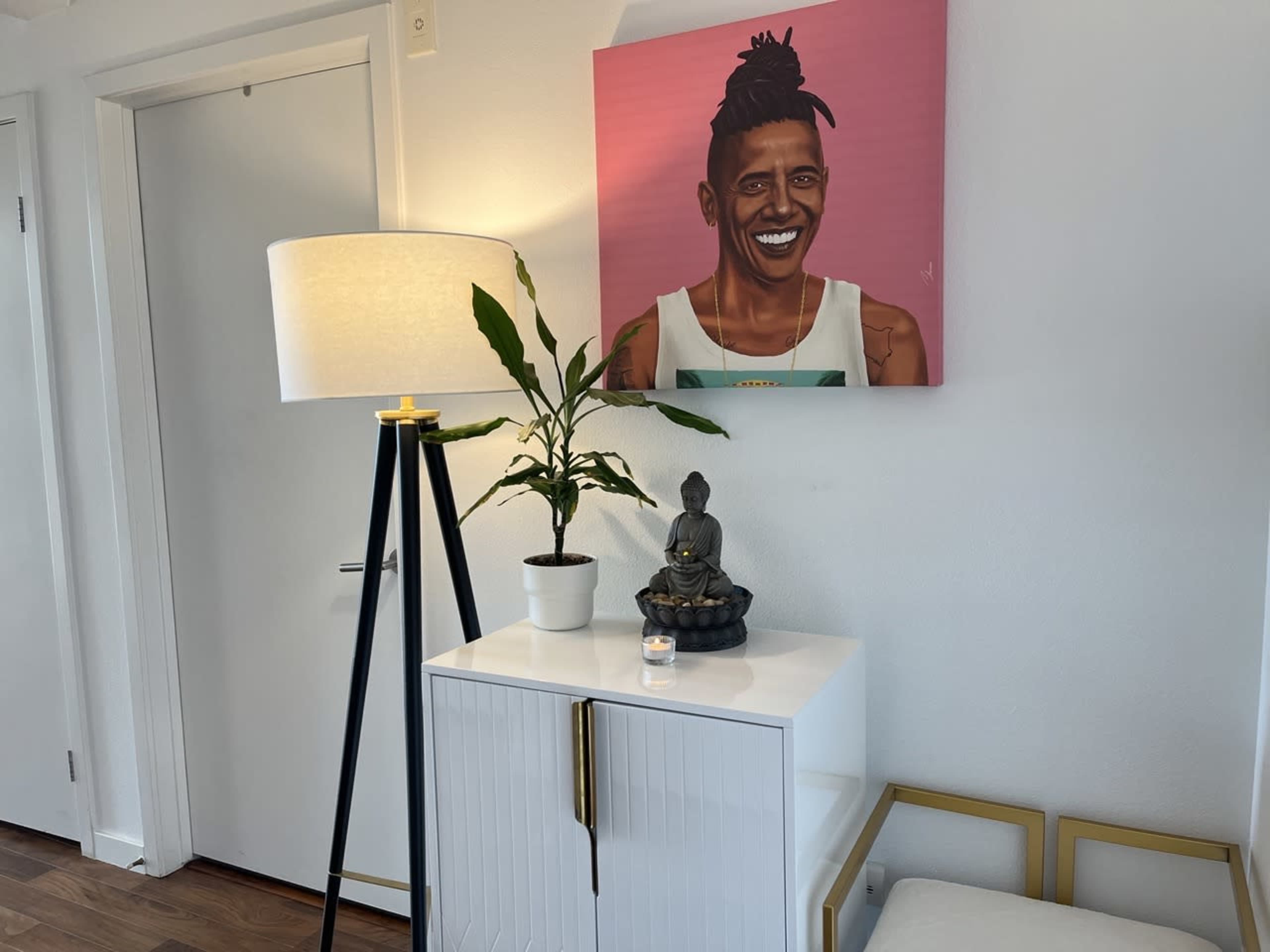 A modern living space features a white side table with a small Buddha statue and a candle, a potted plant beside it, and a lamp with a black stand, all beneath an art piece of a smiling figure on a pink background.