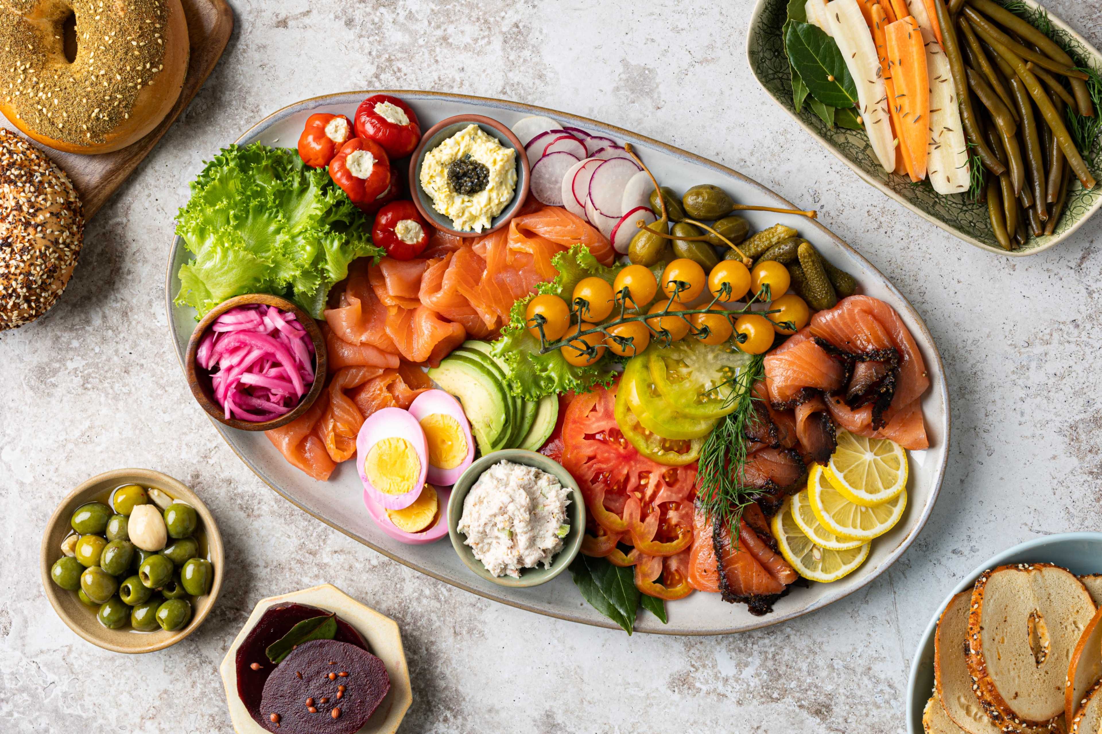 A large platter displays an assortment of smoked salmon, fresh vegetables, pickled items, and various spreads, accompanied by bagels and greens.