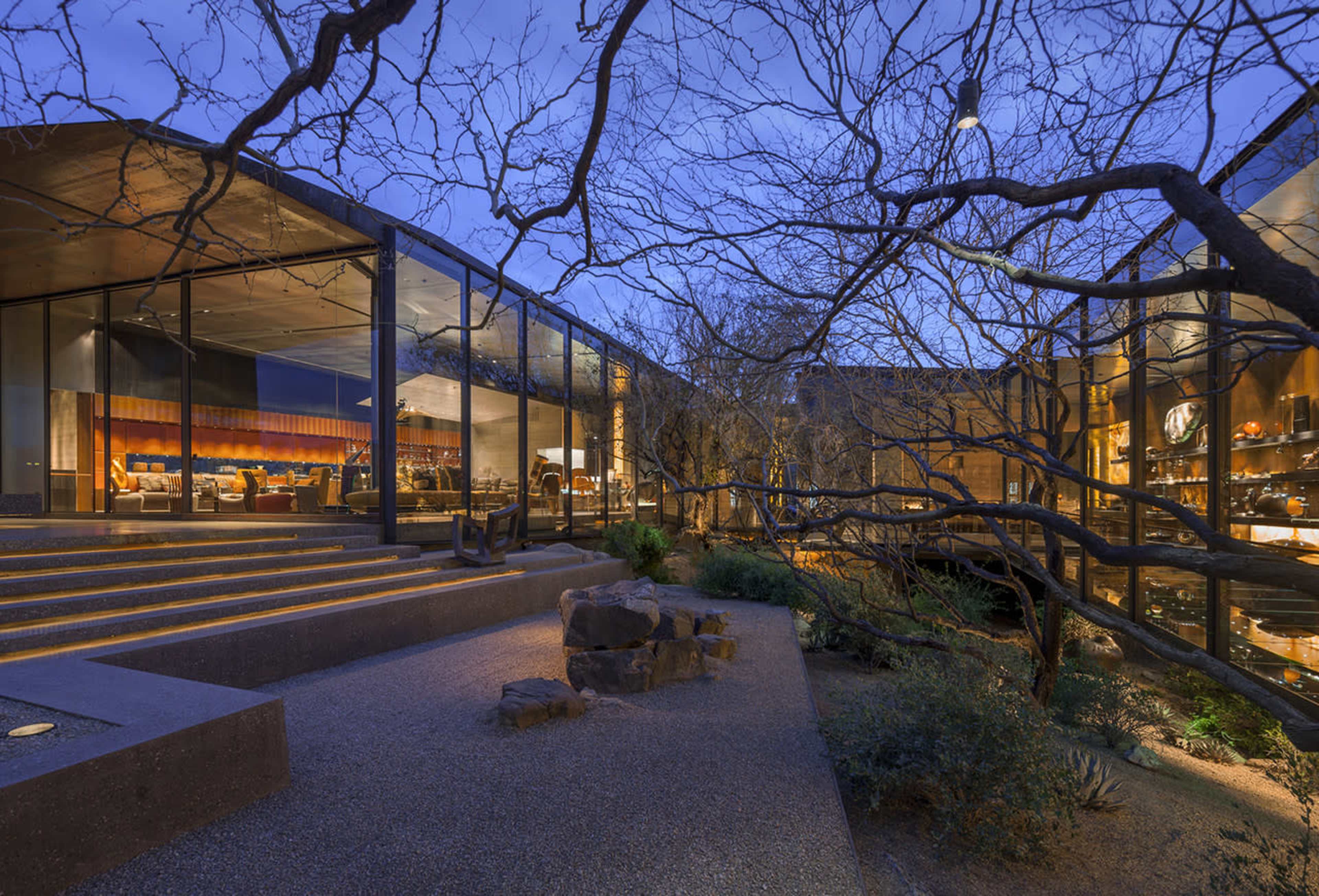 A modern house features large glass walls, surrounded by sparse landscaping and illuminated by soft lighting at dusk.