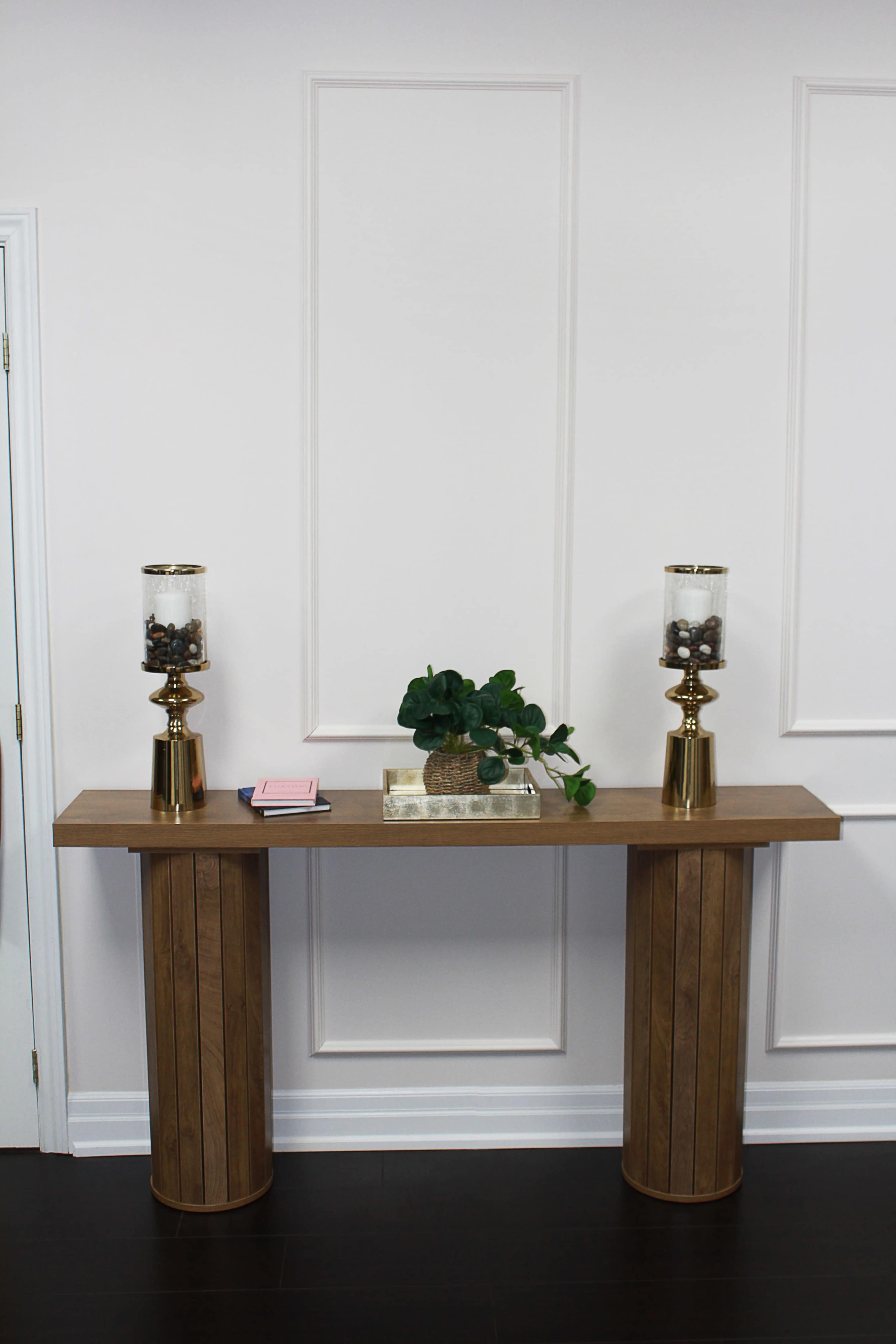 A wooden console table with two cylindrical pedestals supports a decorative arrangement featuring a plant and two glass candle holders.
