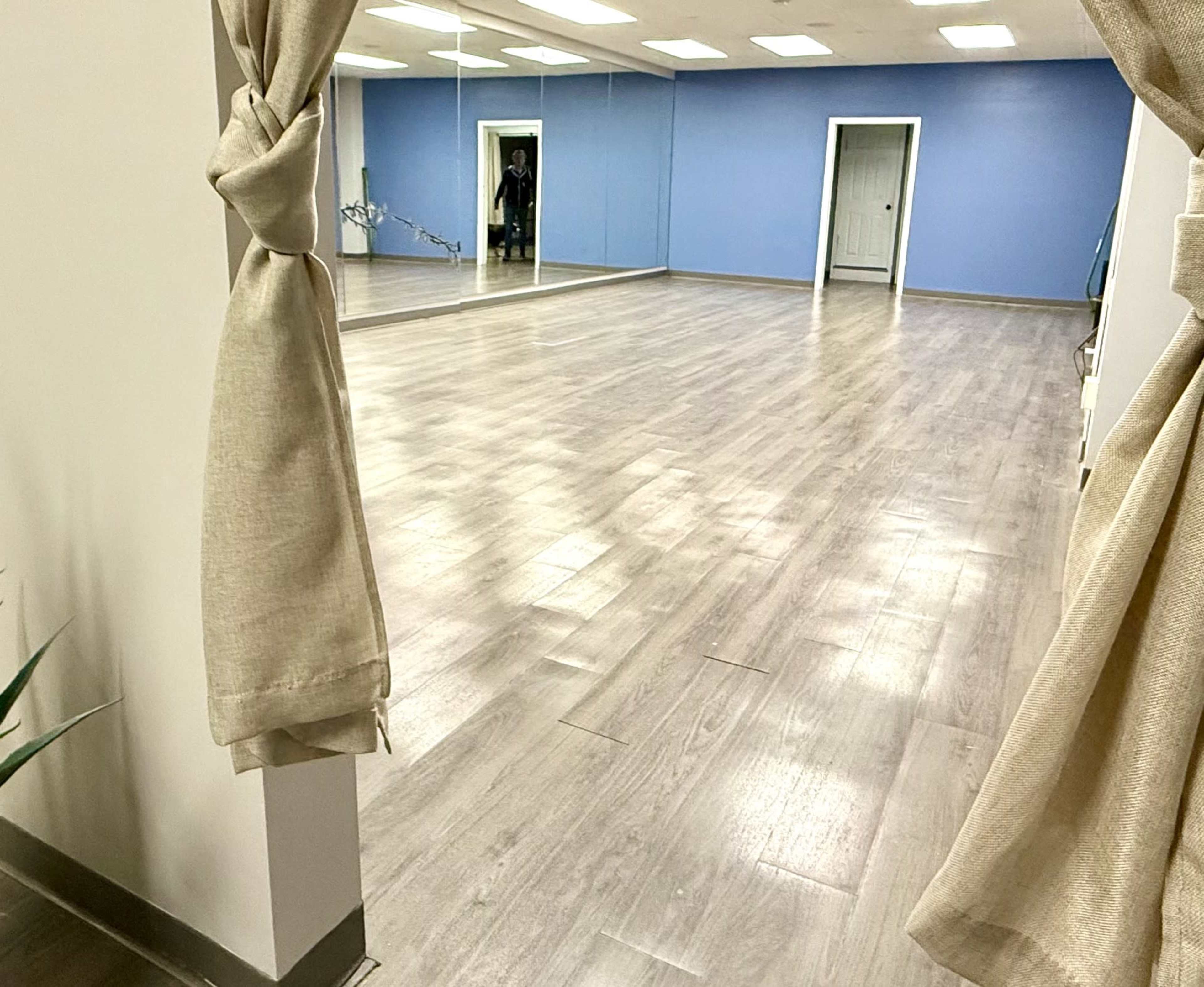 The image shows a spacious, empty room with wooden floors and a blue accent wall, viewed through a curtain tieback.