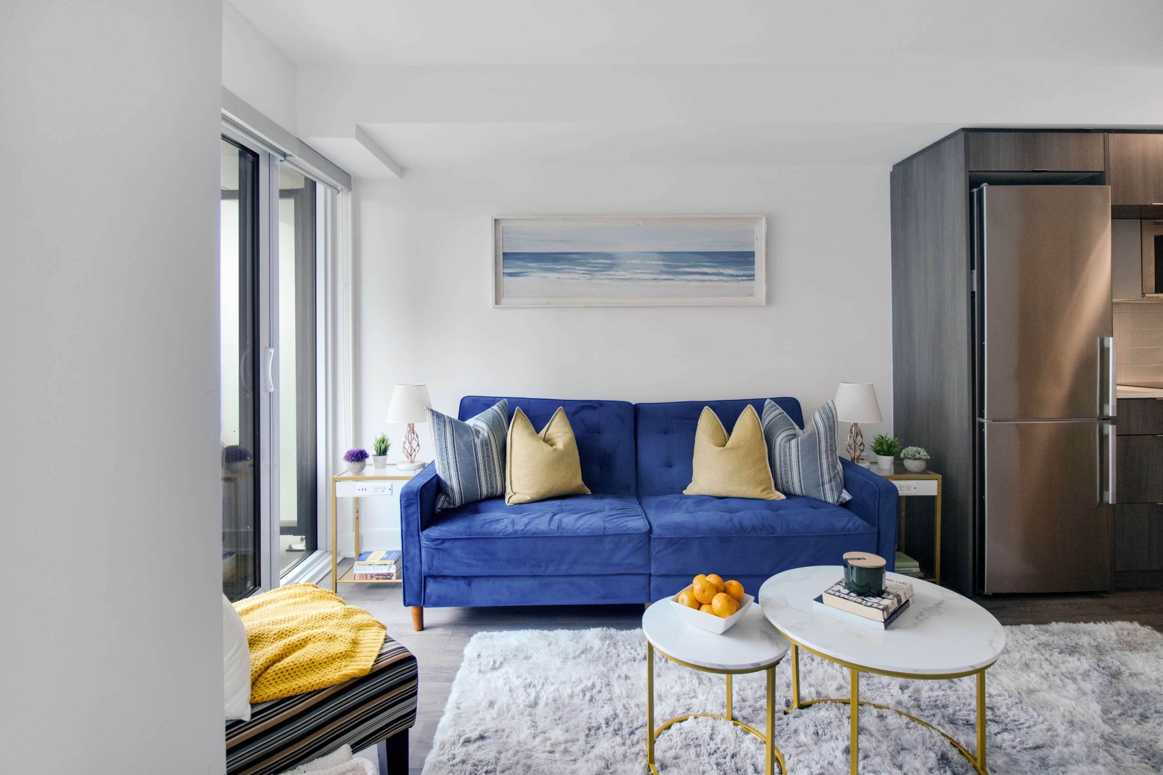 A modern living room with a blue sofa, two side tables, a coffee table with oranges, and a kitchen area in the background.