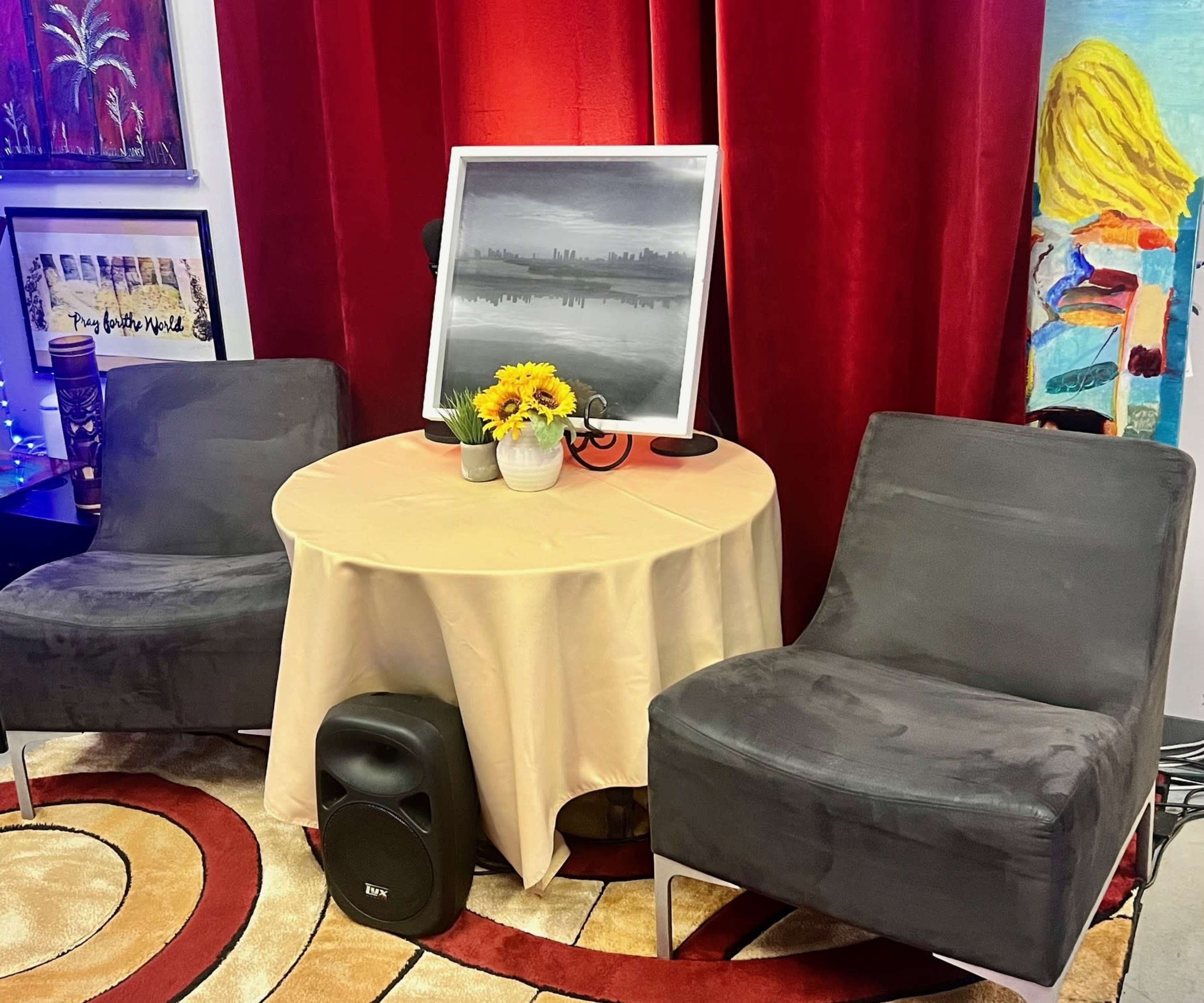 A round table with a beige tablecloth is set between two gray chairs, adorned with a bouquet of sunflowers and a framed painting against a backdrop of red curtains.